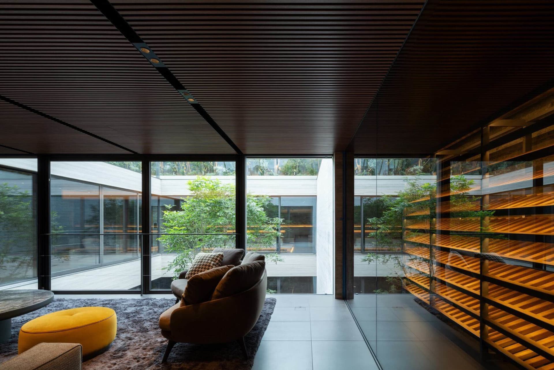 Modern living room featuring a dark wood slatted ceiling, large glass walls overlooking a garden, brown curved sofa, bright yellow ottoman, and illuminated wooden shelving unit.
