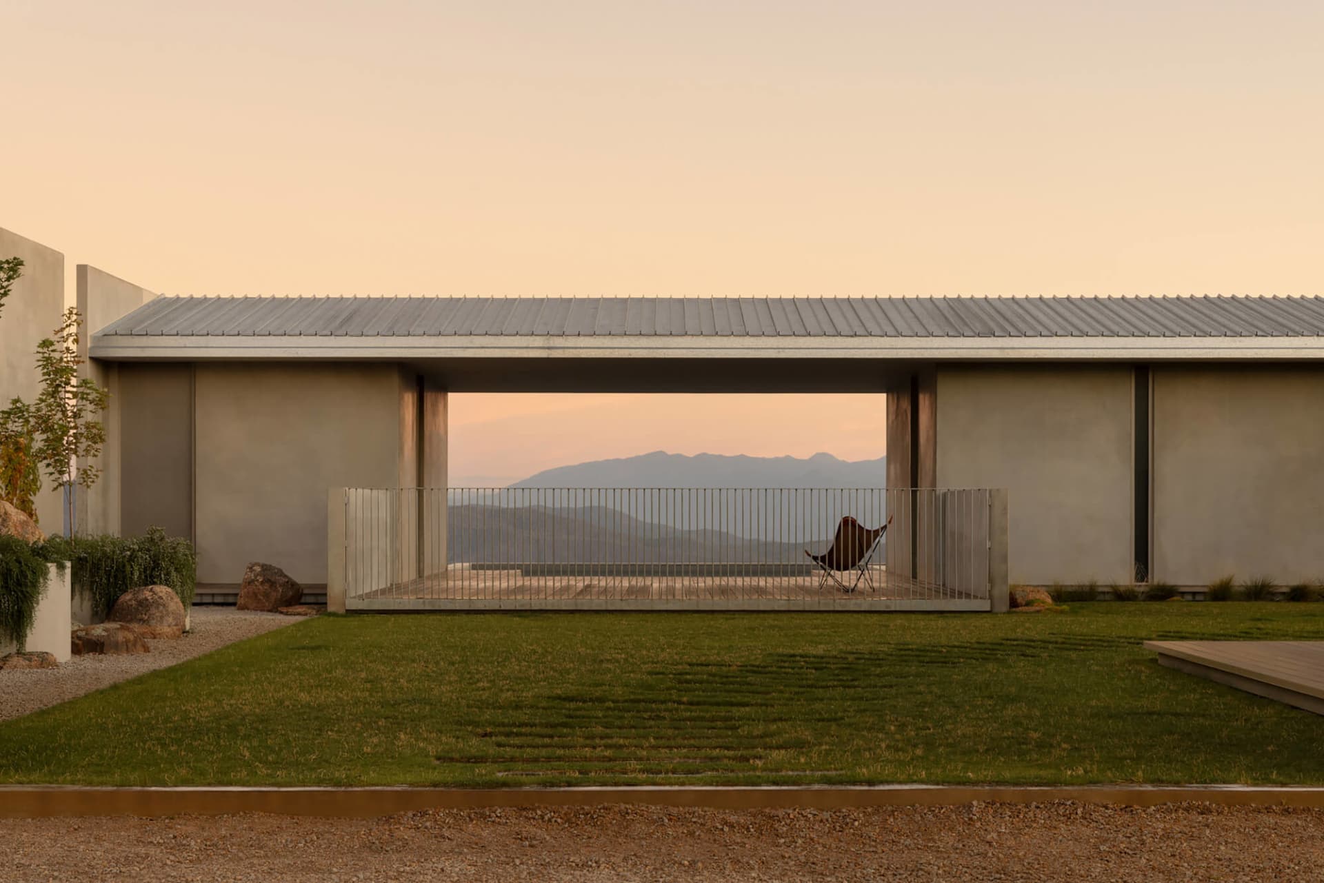 Modern concrete house with open patio, butterfly chair, wooden deck, and distant mountain range at sunset