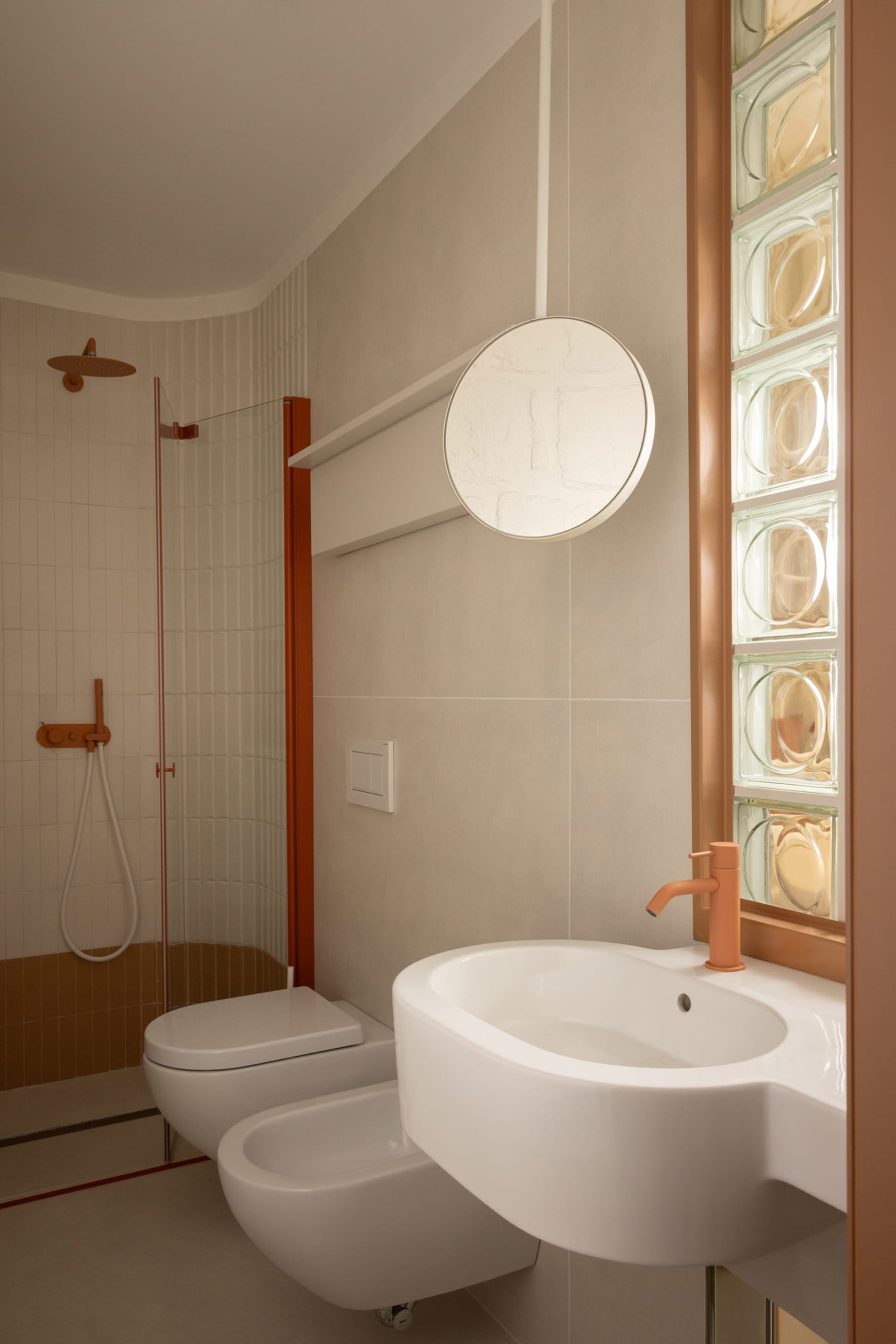 Modern minimalist bathroom with terracotta accents, white wall-hung toilet and bidet, integrated white ceramic sink with matching orange faucet, round mirror, and glass block window.