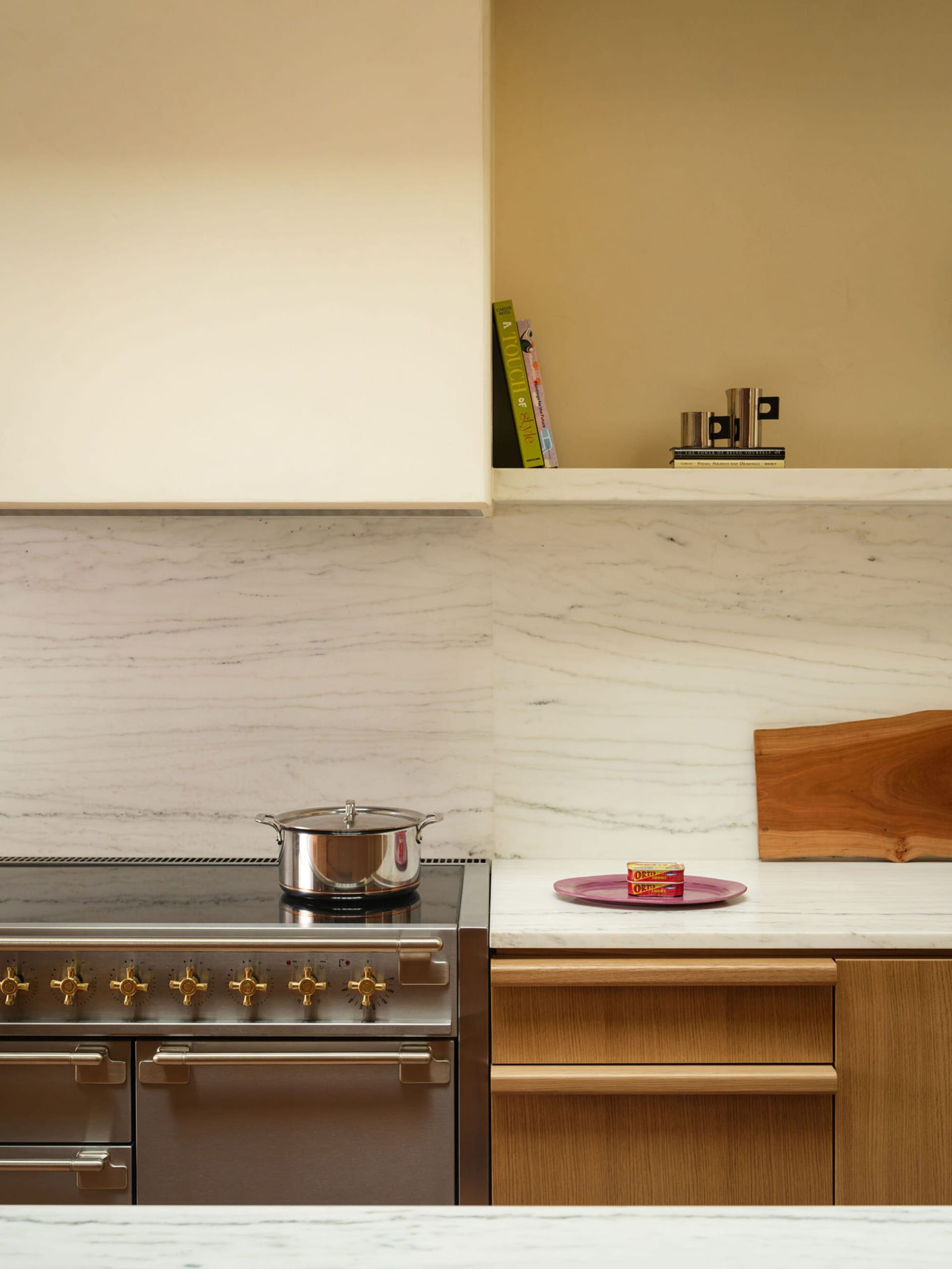 Modern kitchen corner featuring a stainless steel range with brass knobs, white marble backsplash and countertop, light oak cabinetry, and a wooden cutting board with colorful cookbooks on an upper shelf.