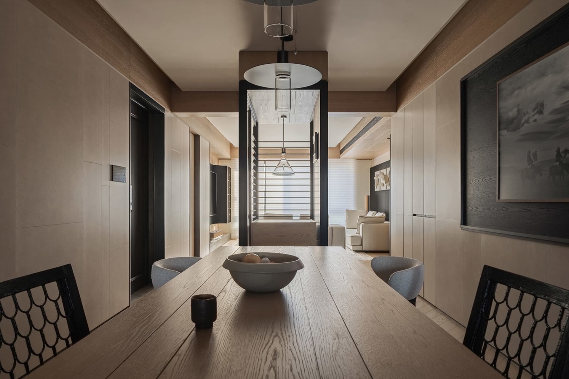 Modern minimalist dining room featuring a long light oak table, black lattice-back chairs, wood panel walls, and a view through to a living area.