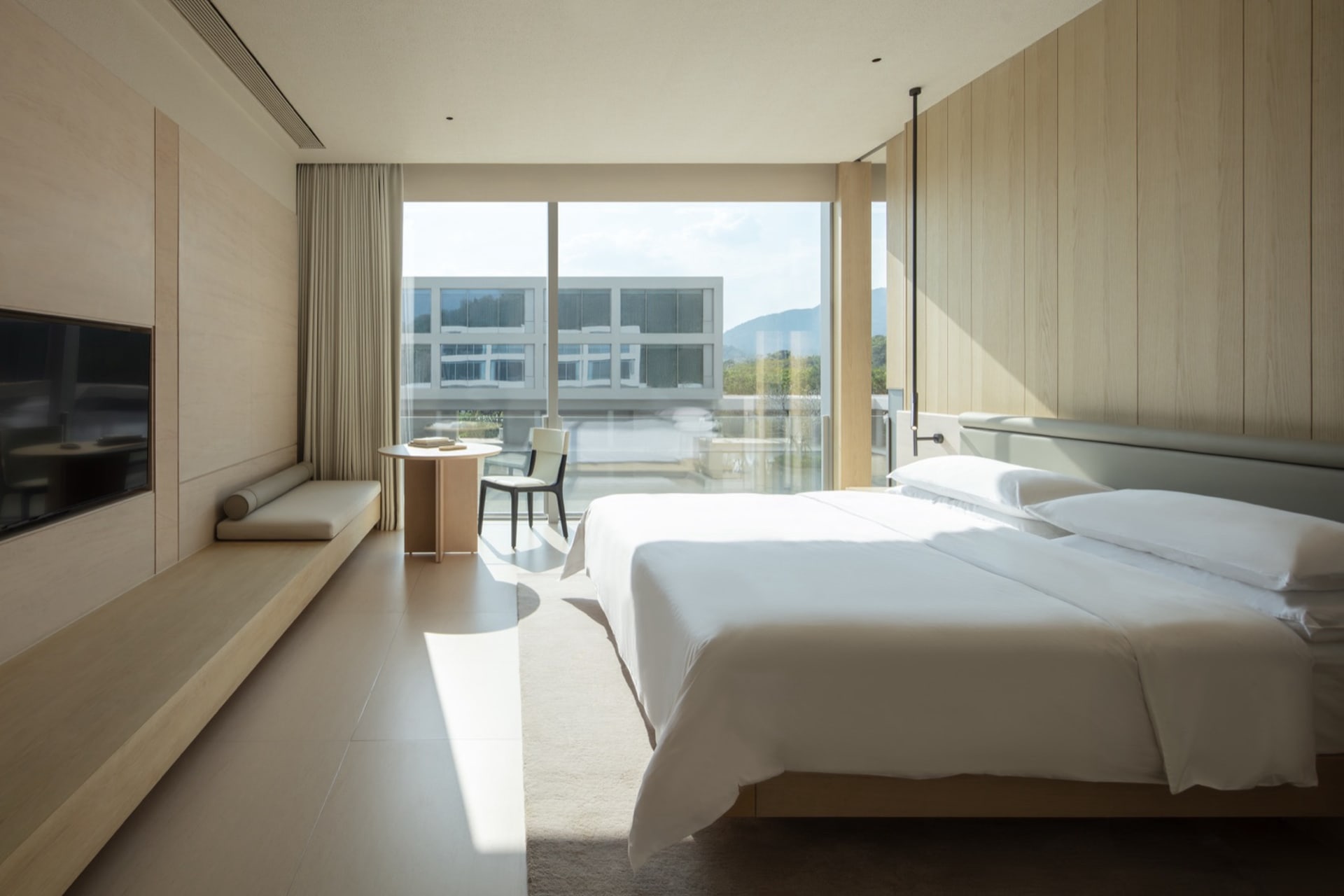 Modern minimalist hotel bedroom featuring light oak wood paneling, a large platform bed with white linens, built-in window seating, and a stunning mountain view through floor-to-ceiling glass doors.