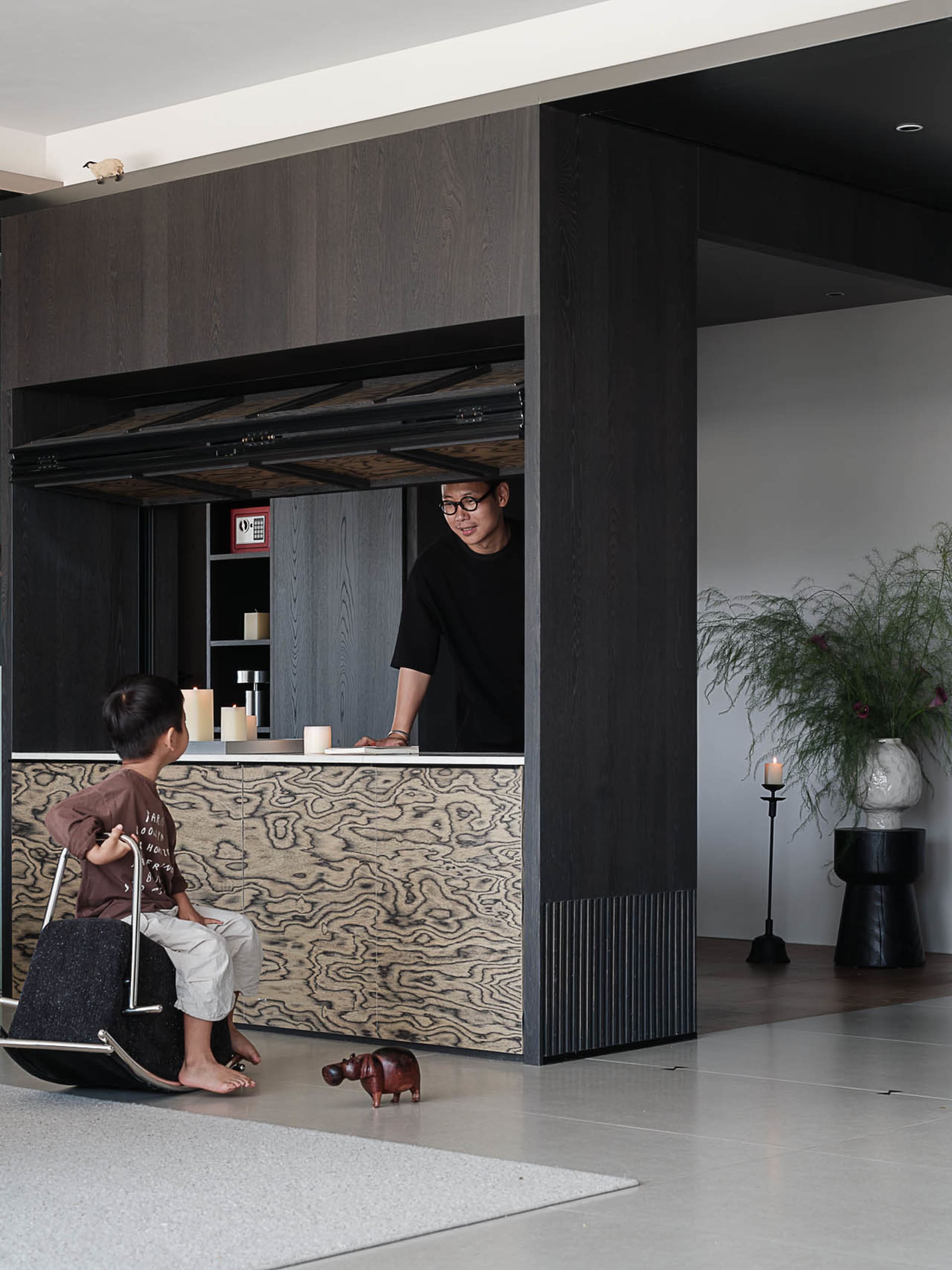 Modern dark-toned living room featuring a large burl wood patterned pass-through counter, a man standing behind the counter and a young boy sitting on a rocking chair in front, with a potted plant and candle on the right side.