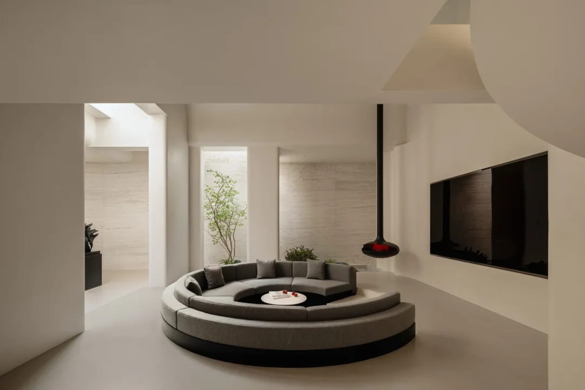 Modern minimalist living room featuring a large grey curved modular sofa on a circular platform, limestone walls, high ceilings with skylights, and a black pendant fireplace.