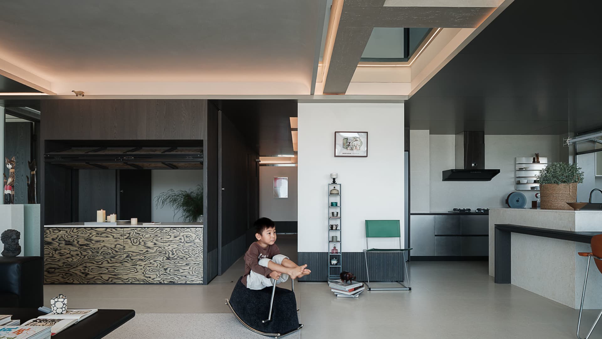 Modern open-plan living and kitchen area with dark wood grain feature wall, concrete ceiling details, and a child playing on a rocking horse.