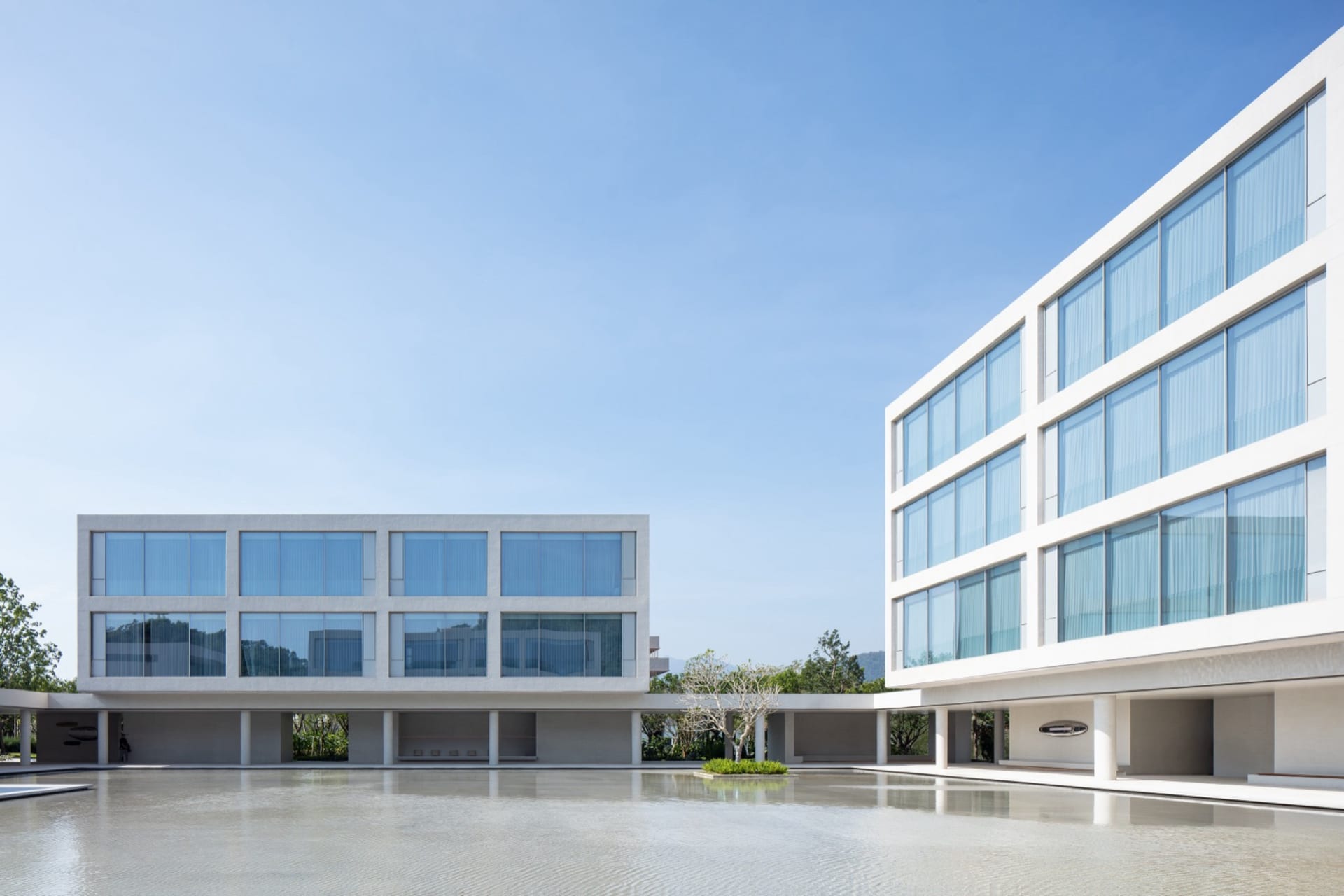 Modern two-story concrete and glass courtyard house with a large reflecting pool in the foreground under a bright blue sky.