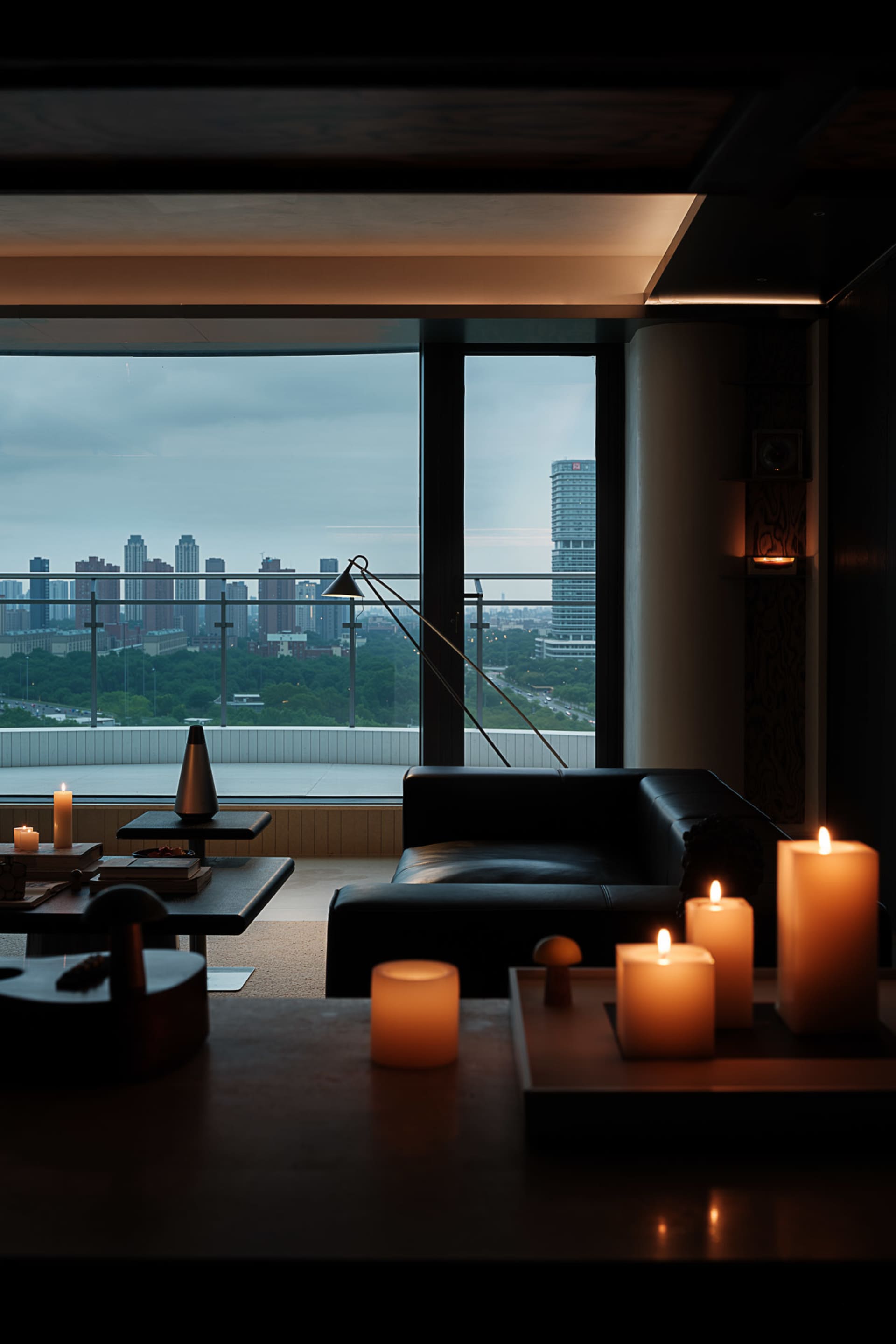 Modern living room interior at night featuring a black leather sofa, multiple lit candles, minimalist dark wood furniture, and large floor-to-ceiling windows overlooking a city skyline with buildings and greenery.