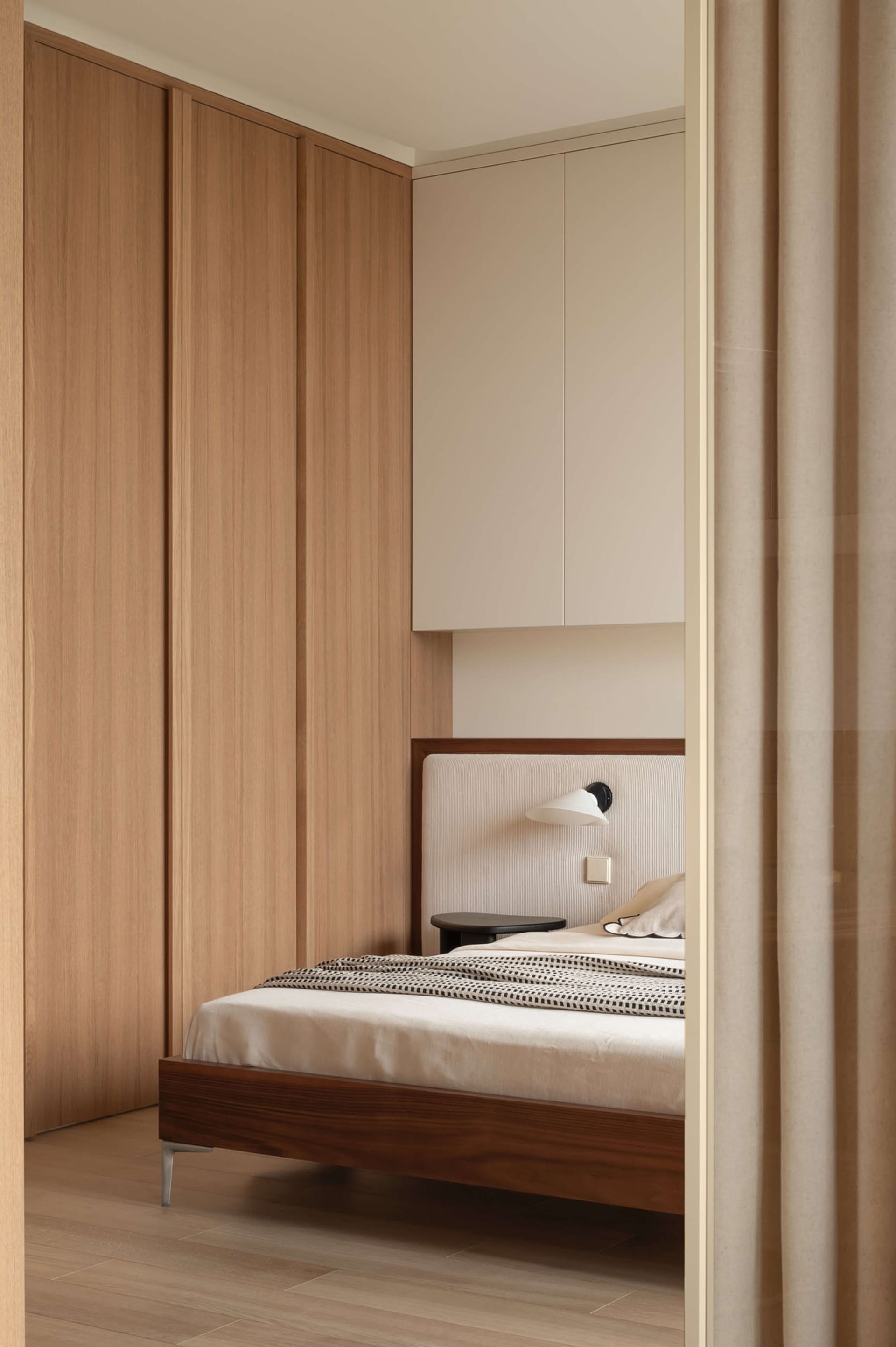 Modern minimalist bedroom featuring a dark walnut platform bed with a ribbed upholstered headboard, built-in light wood wardrobes, wall-mounted reading lamp, and neutral beige curtains.