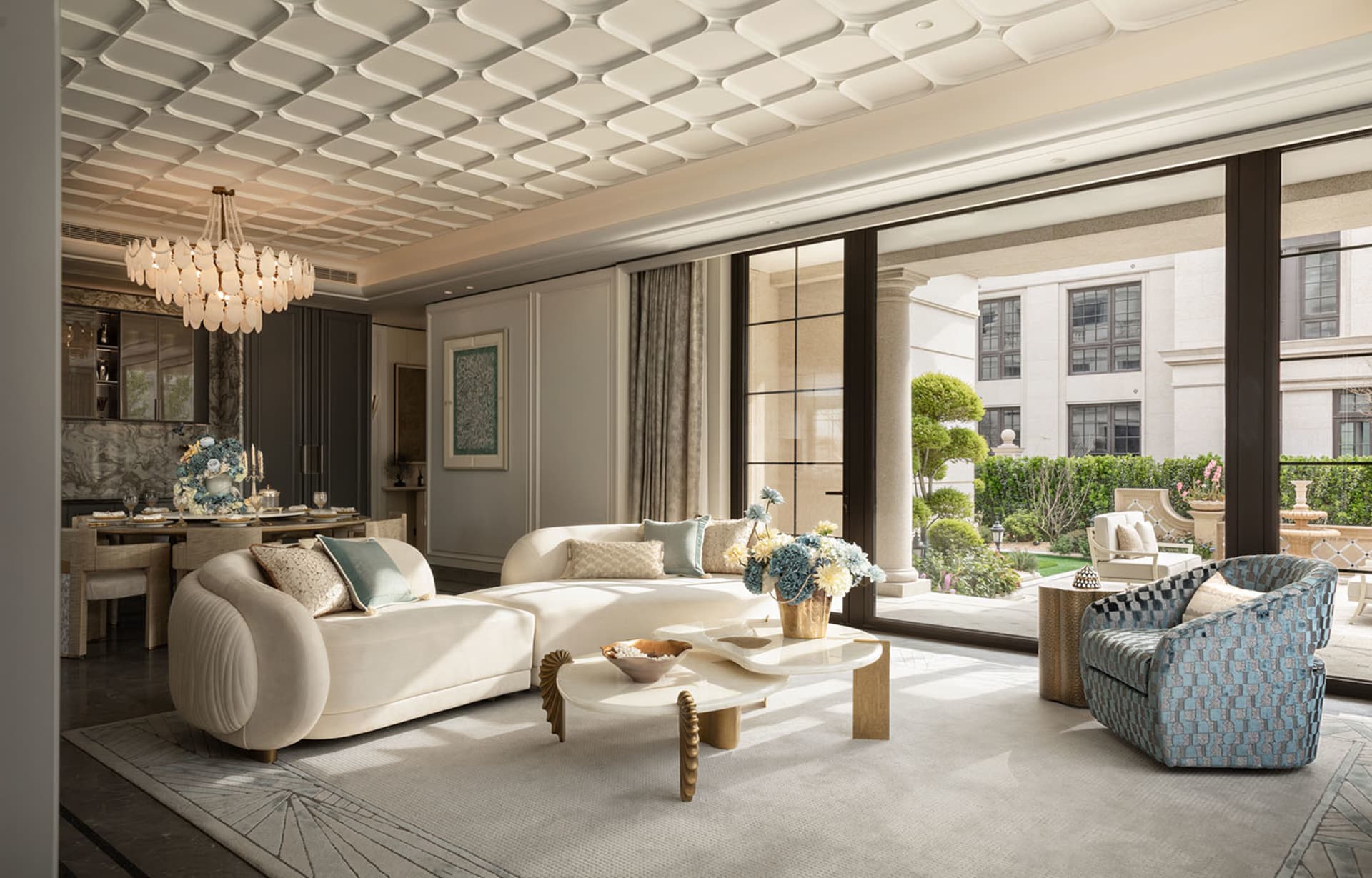 Bright and airy living room featuring a curved cream sofa, blue patterned armchair, organic-shaped marble coffee table, and large glass doors opening to a landscaped garden.