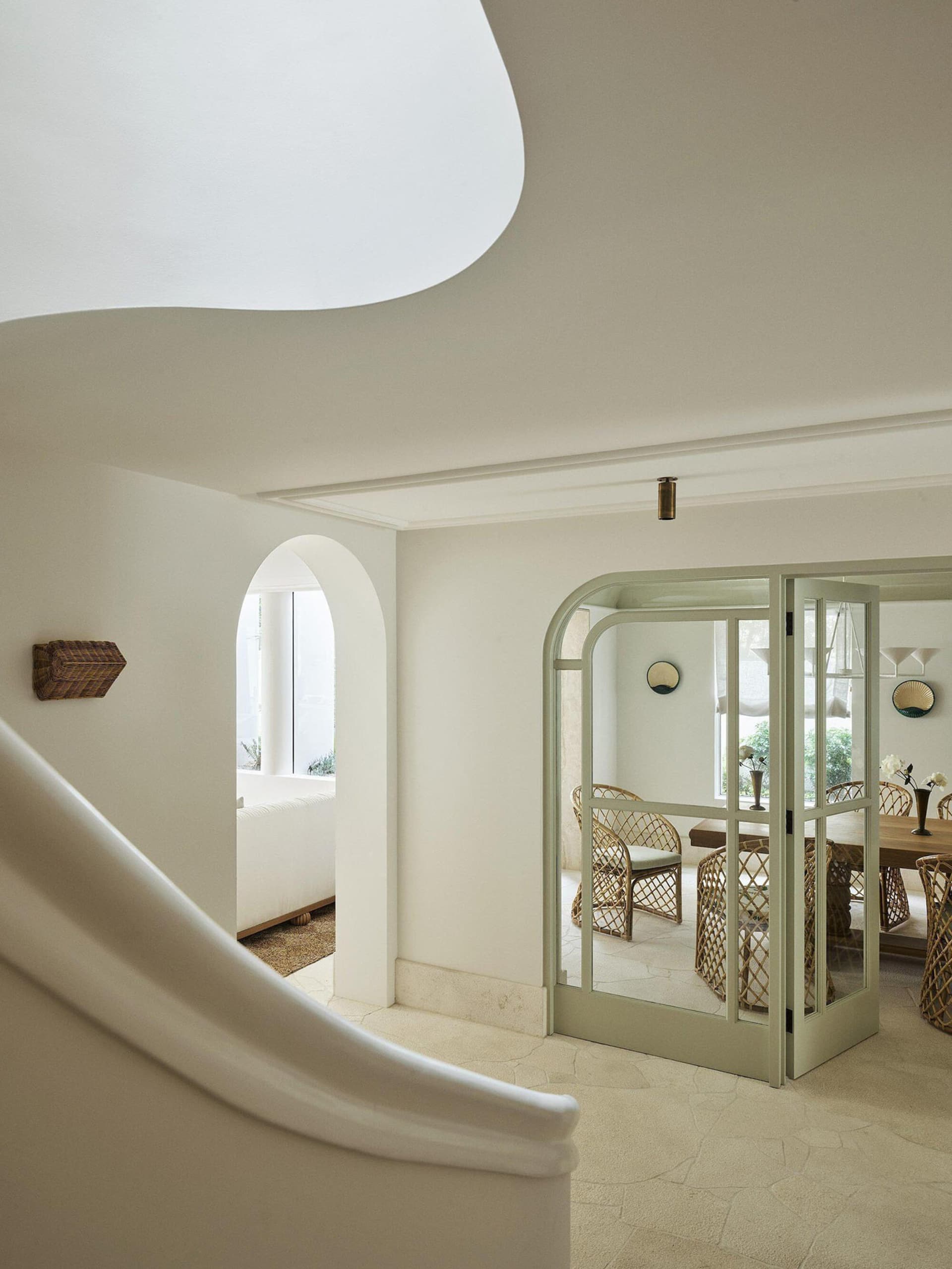 View from a curved staircase into a bright dining room with sage green arched French doors, rattan dining chairs, and white plaster walls.