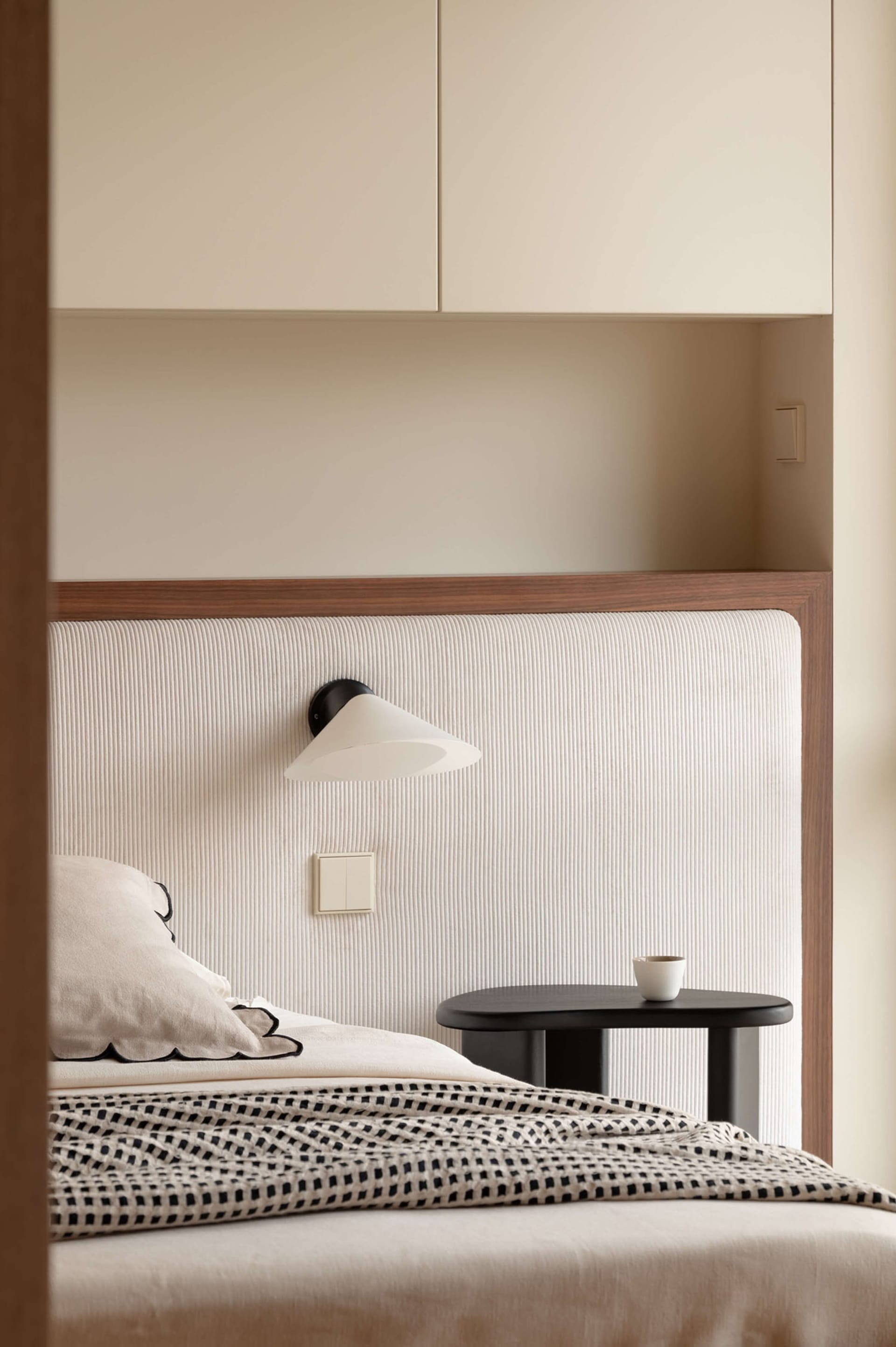 Modern minimalist bedroom with a cream-colored vertically ribbed fabric headboard, black cone-shaped wall sconce, dark oval bedside table, and neutral bedding with black-and-white checkered throw.