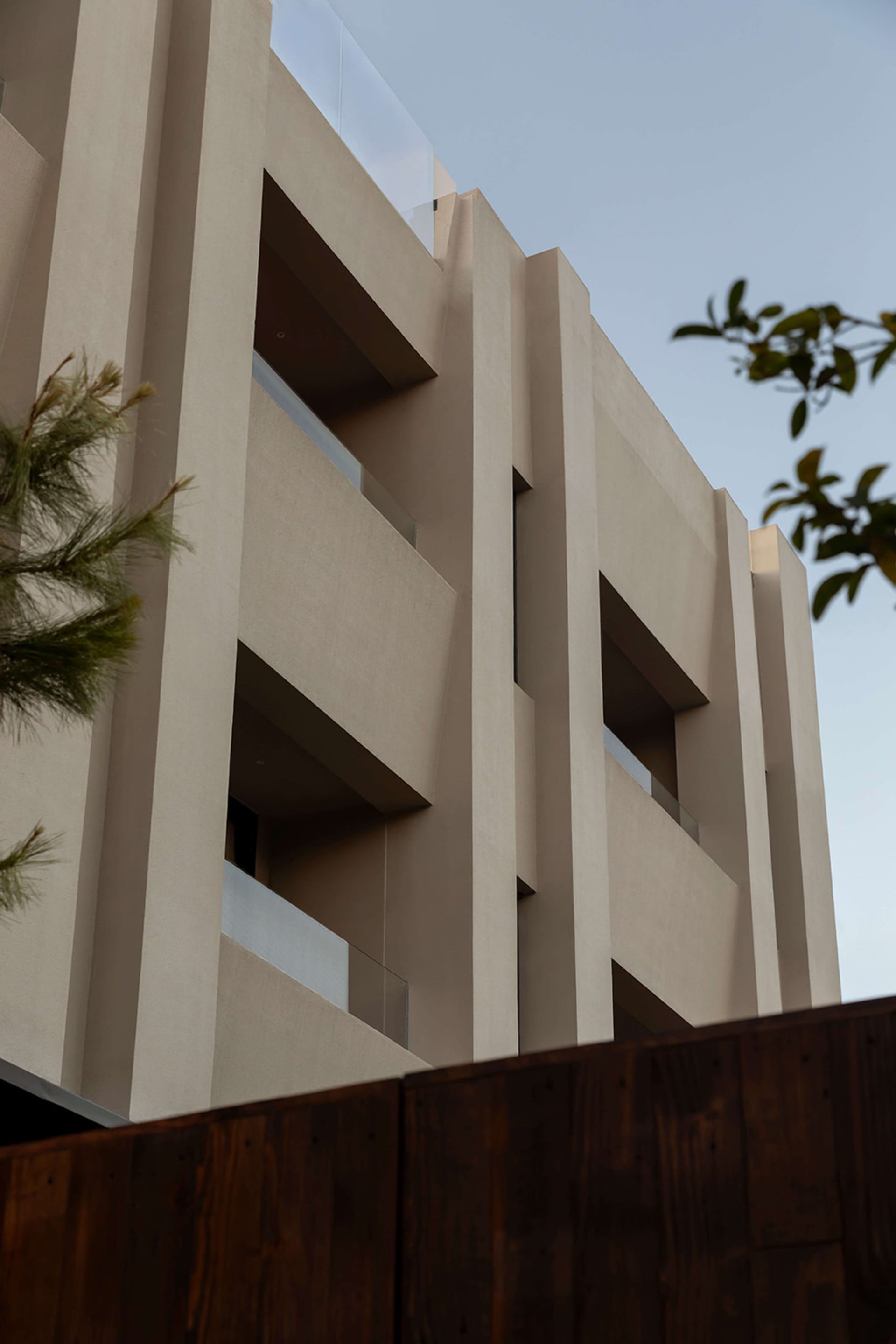Low-angle view of a modern multi-story concrete building featuring protruding structural elements, recessed balconies with glass railings, and a dark wooden fence in the foreground with tree branches on the sides.