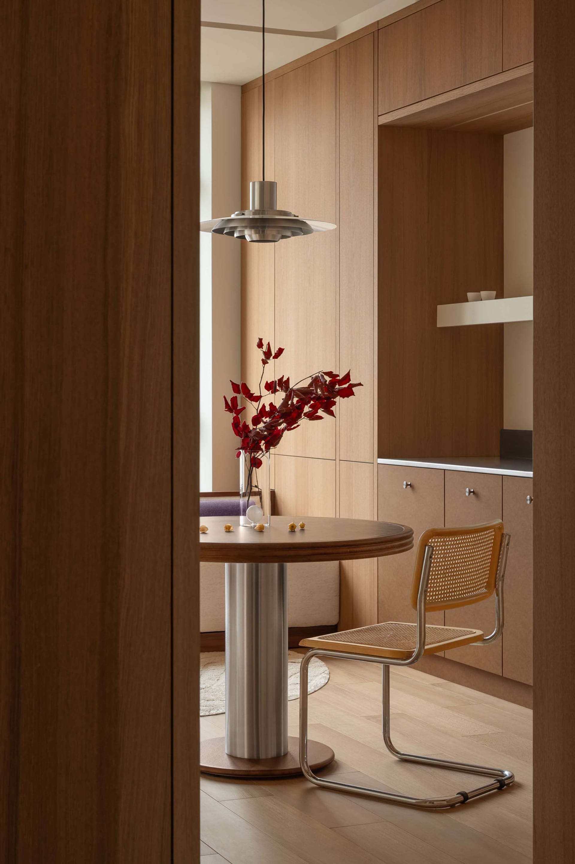Modern minimalist breakfast nook featuring light walnut cabinetry, a round pedestal dining table, a cane and chrome chair, and a Louis Poulsen PH 5 pendant lamp with red floral arrangement.