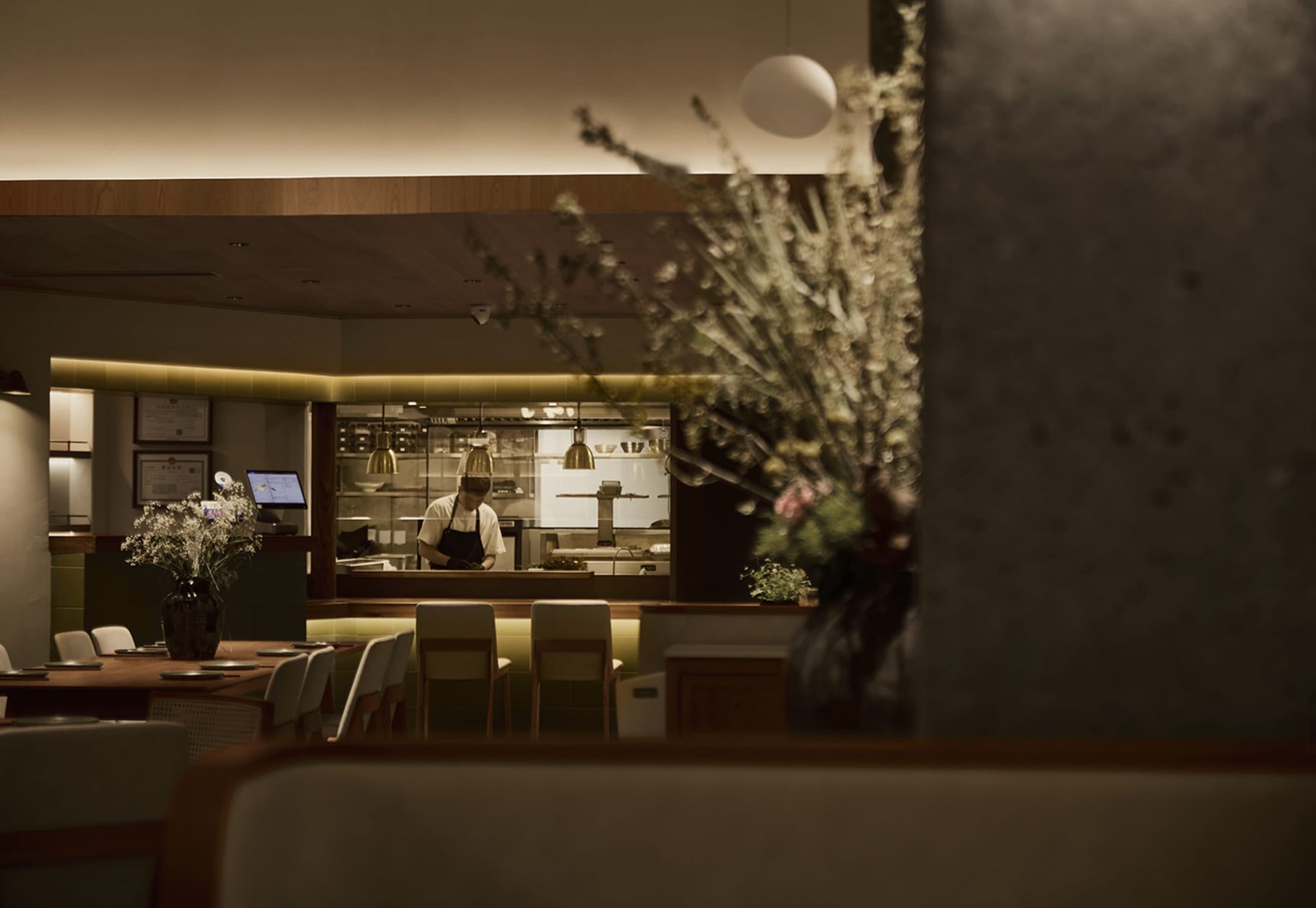 View from a dining table looking past a foreground vase of dried flowers toward an open kitchen where a chef is working under warm pendant lights, with wooden tables, upholstered chairs, and concrete walls.