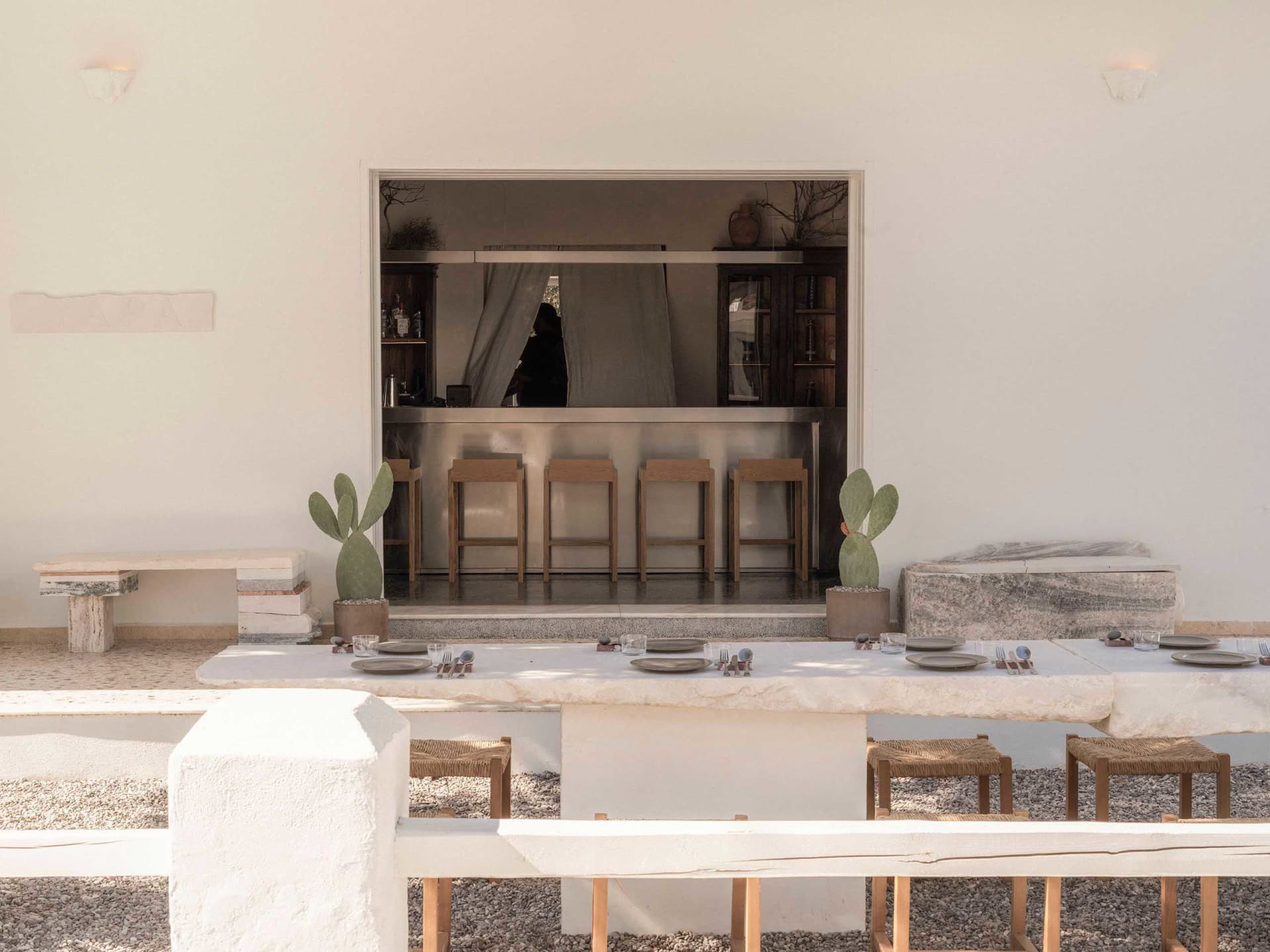 Outdoor dining area with a long travertine table, woven stools, potted cacti, and a view through a large opening to an indoor bar with wooden stools.