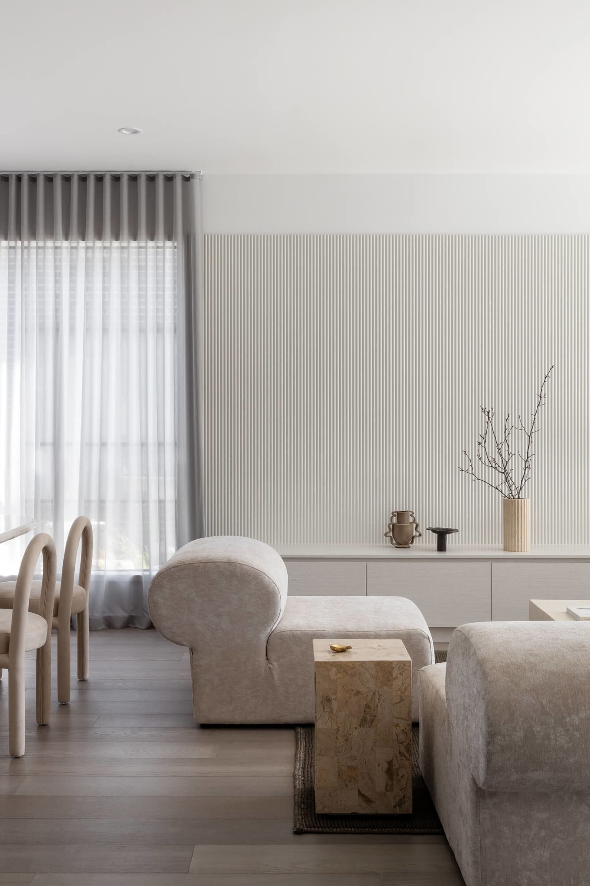Modern minimalist living room featuring a curved bouclé lounge chair, a travertine block side table, fluted wall paneling, and neutral beige tones.