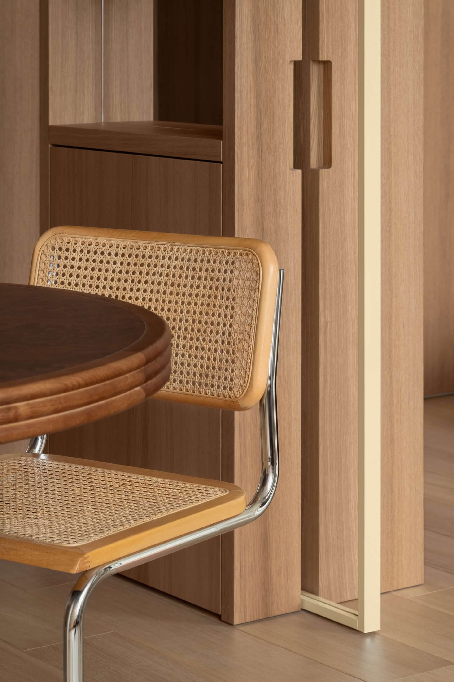 Cane and walnut dining chair with chrome frame next to a round walnut dining table in a wood-paneled dining room