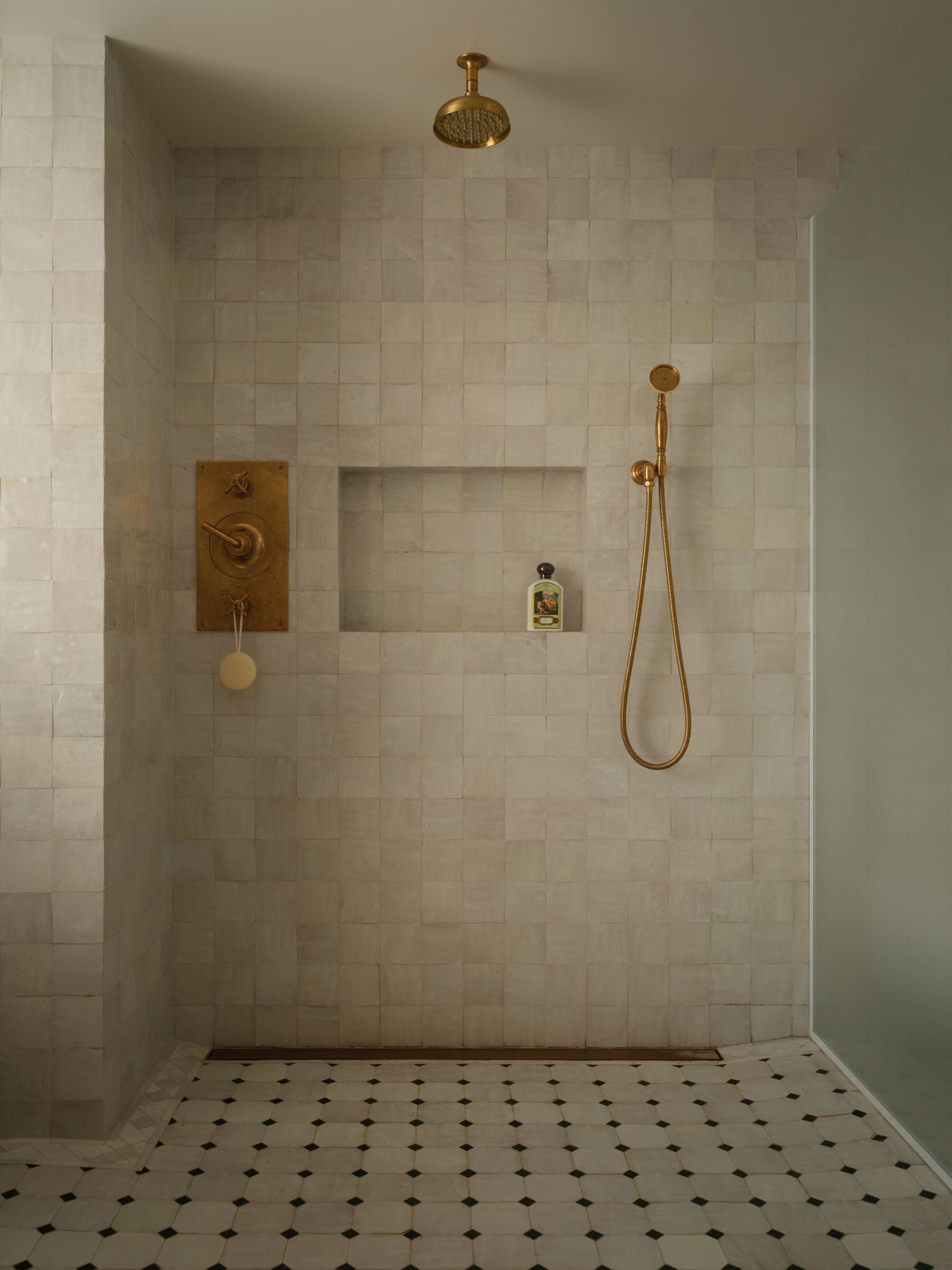 Minimalist shower space with off-white zellige tiled walls, a recessed niche holding a bath product, unlacquered brass shower head and mixer, and geometric black-and-white mosaic floor tiles.