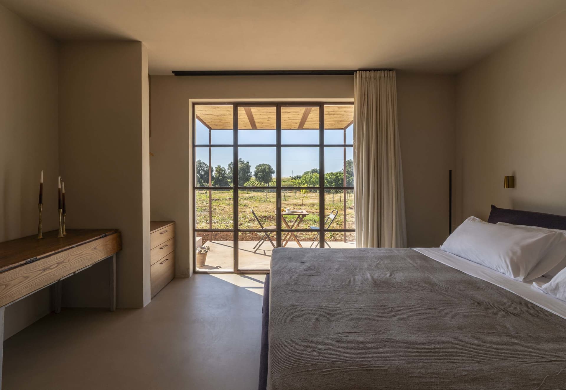Modern minimalist bedroom featuring a large bed with grey bedding, wooden console desk, and floor-to-ceiling glass doors opening to a vineyard terrace.