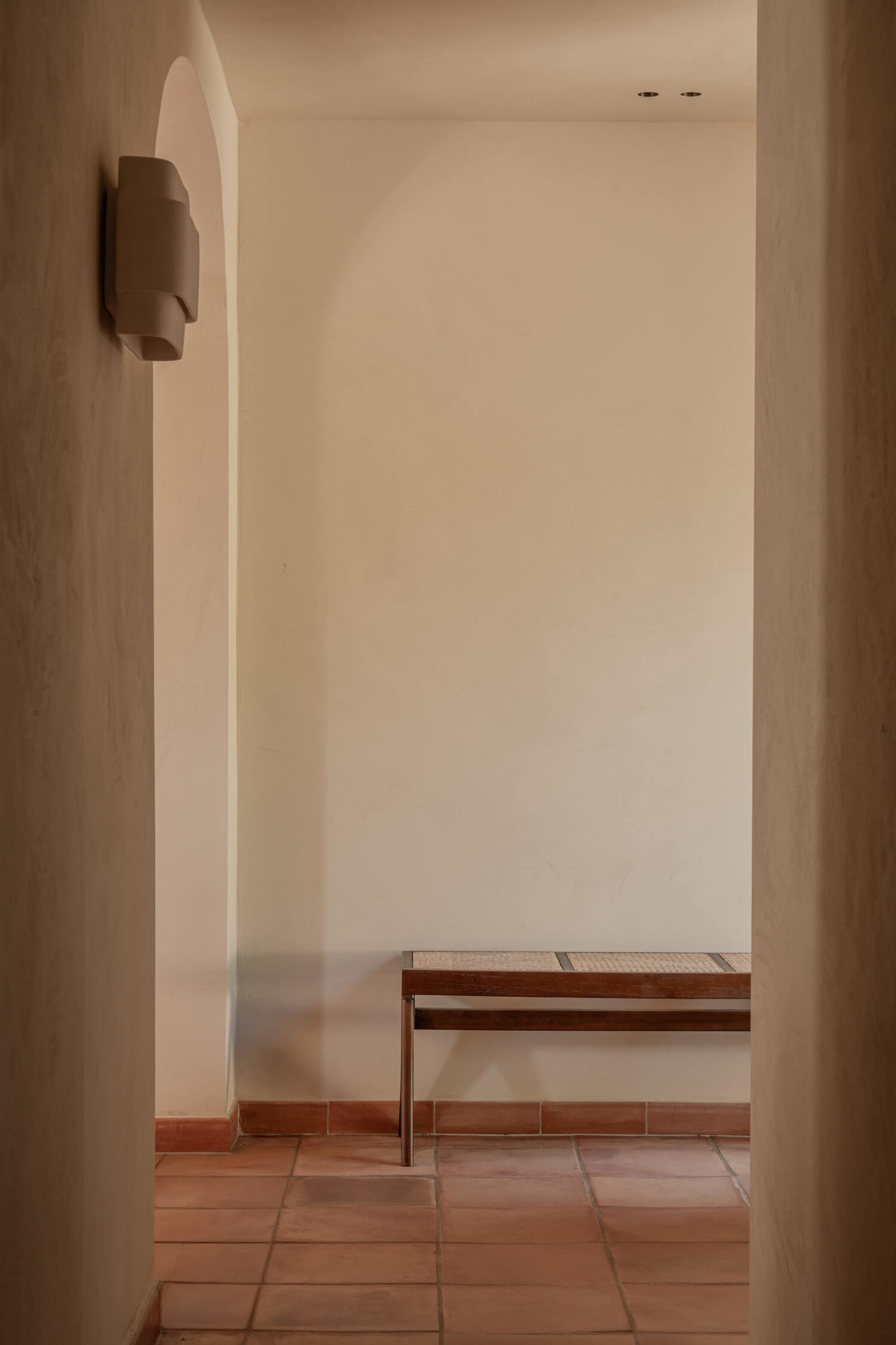 Minimalist entryway featuring terracotta floor tiles, a wooden bench with woven seat, and a sculptural wall sconce under an arched alcove.