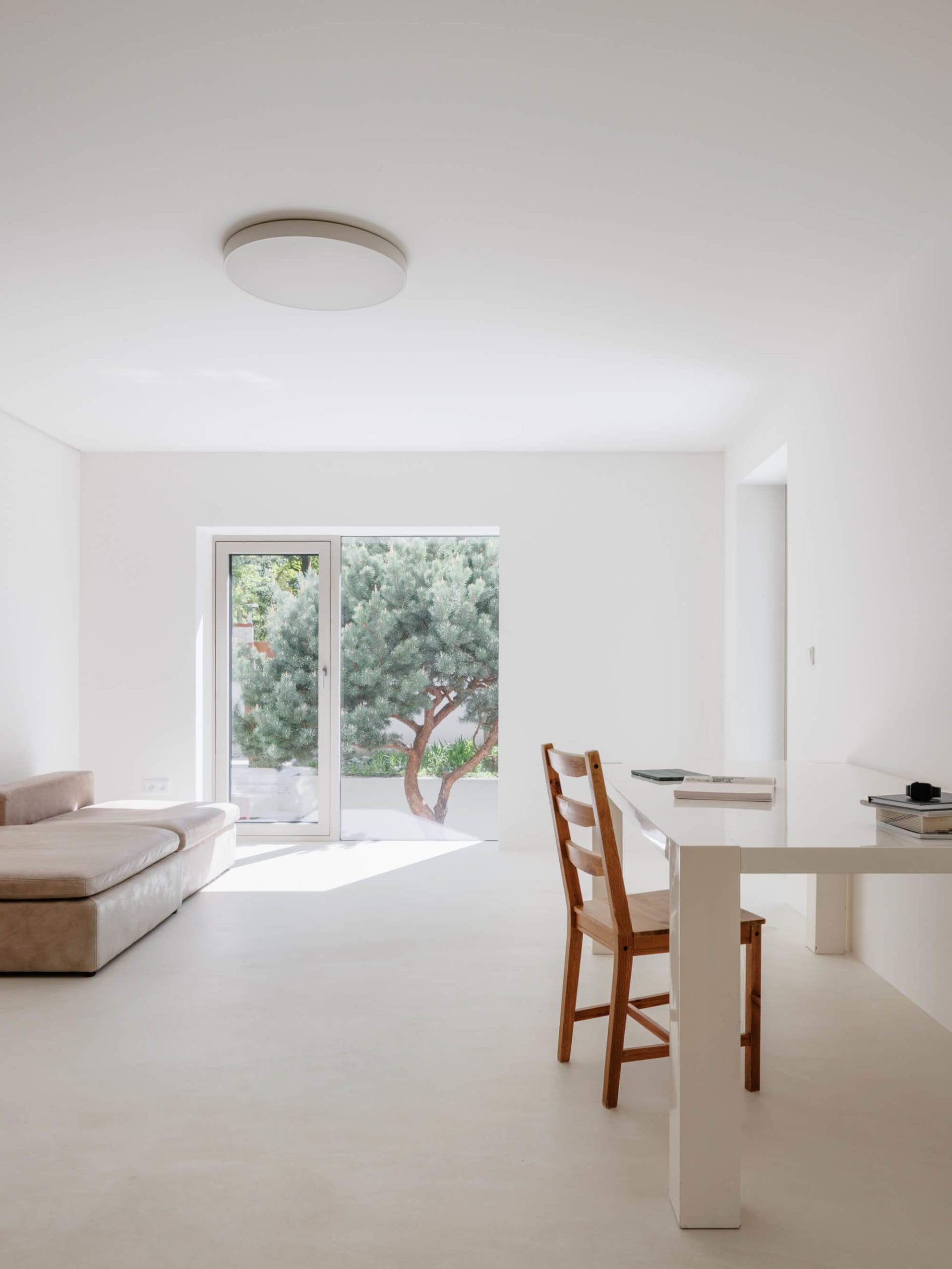 A bright, minimalist dining room with white walls and floor, a wooden dining chair, a white table, and a large glass door opening to a green tree outside.