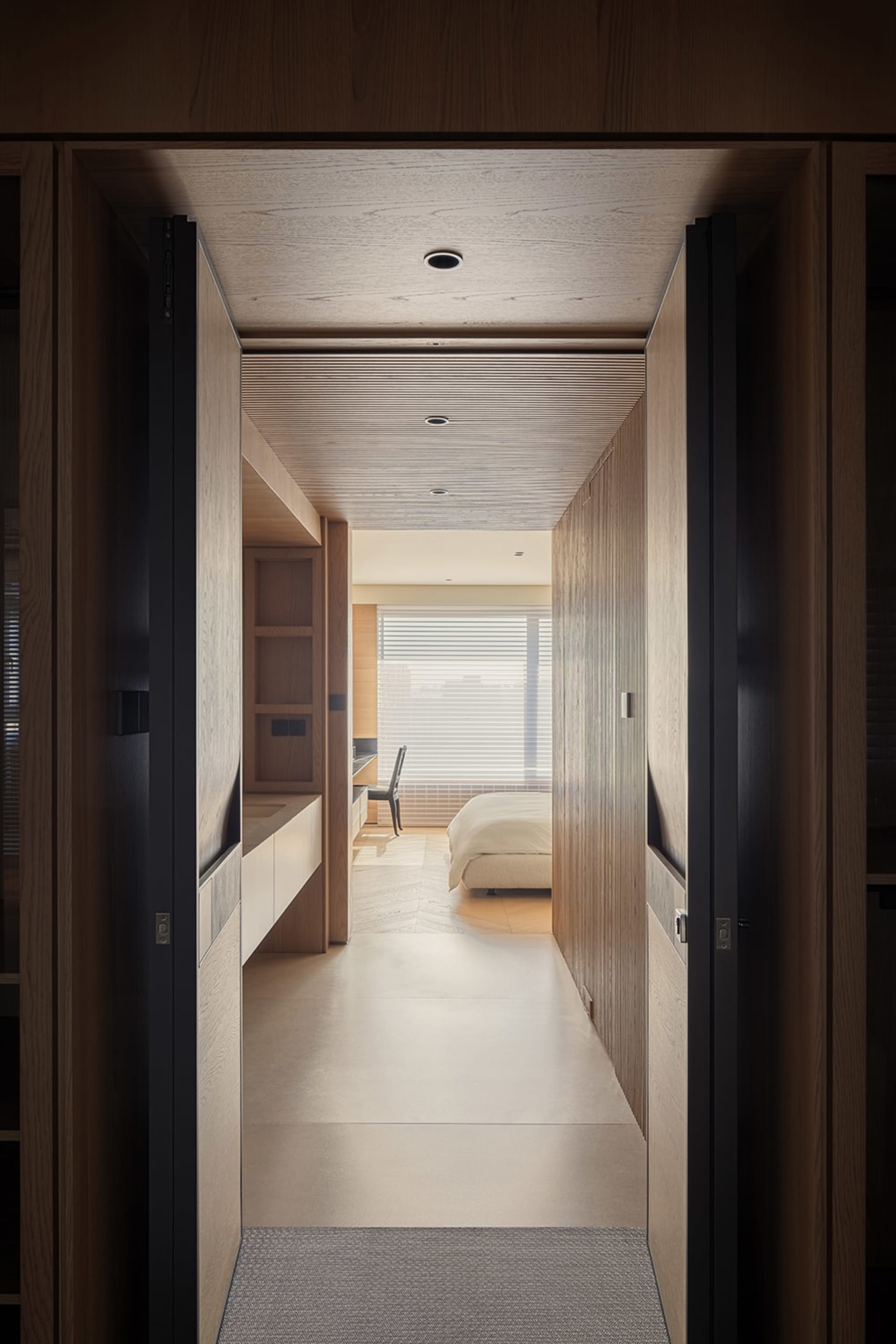View from a doorway looking down a minimalist hallway with oak wood walls, built-in storage, and a wood slat ceiling leading to a bright bedroom with a large window.
