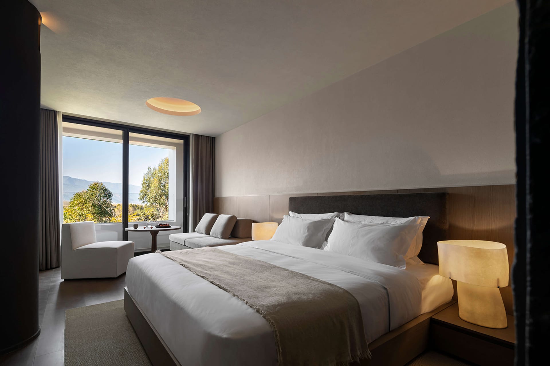 Modern hotel bedroom with large bed dressed in white linens, dark wood headboard, upholstered sofa, and large windows overlooking green mountains and trees.