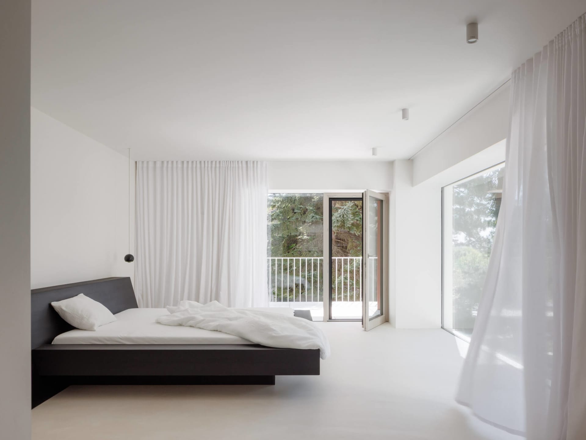 Modern minimalist bedroom with a dark wood floating platform bed, white bedding, sheer white curtains, and large glass doors opening to a balcony with greenery.