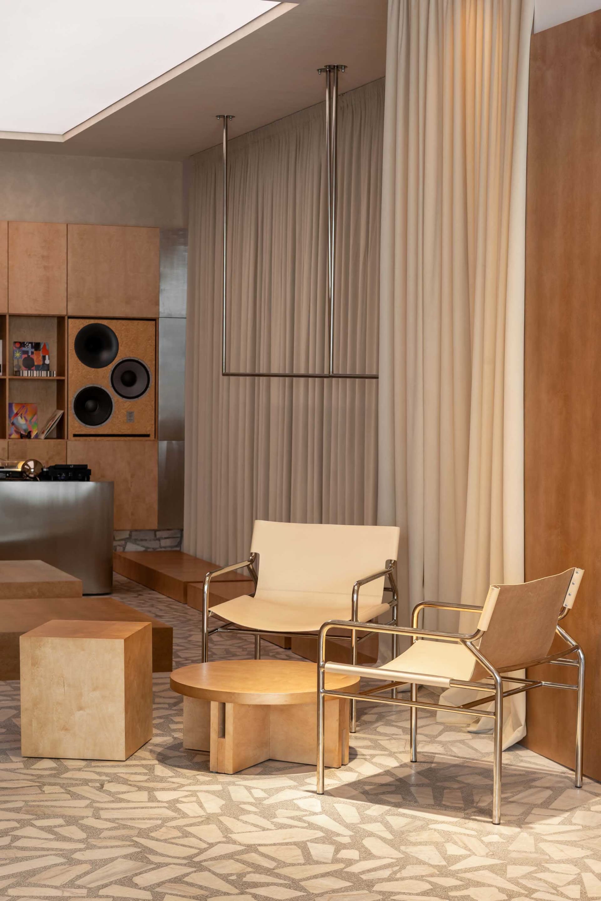 Modern living room featuring two cantilevered lounge chairs with chrome frames and cream upholstery, a round travertine coffee table, stone flooring, and a built-in wooden audio cabinet with speakers.