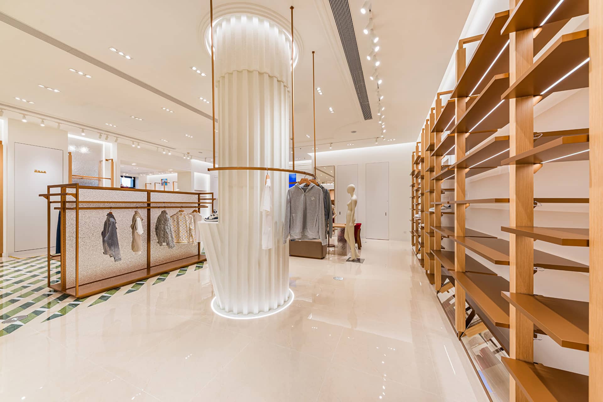 Modern minimalist clothing boutique interior with tall oak wood shelving units on the right, a large white pleated central column with integrated lighting, polished light-colored tile flooring, and clothing racks on the left.
