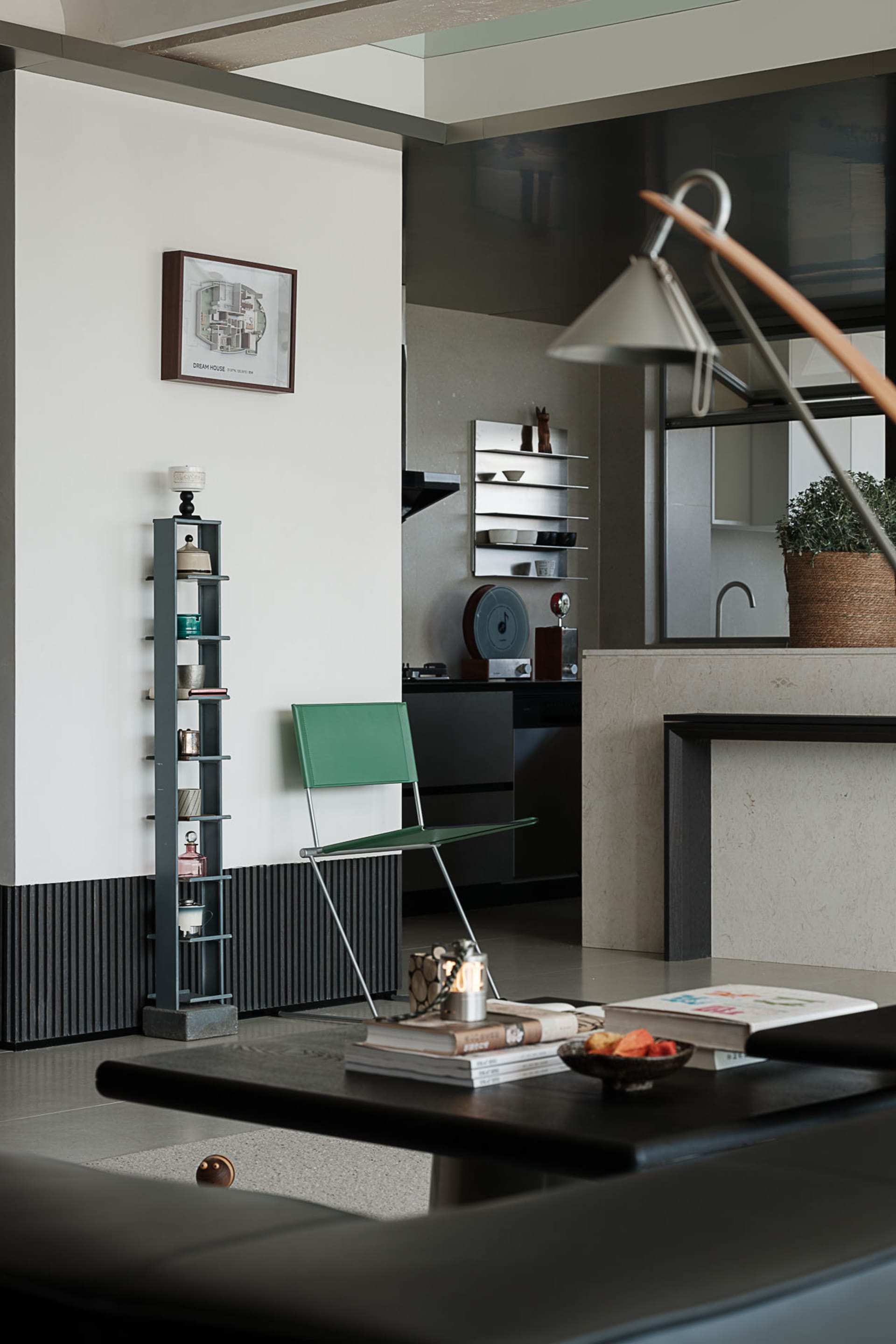 Modern minimalist living room featuring an emerald green chair, dark grey tall shelving unit, concrete kitchen island, and warm lighting from a sculptural floor lamp.