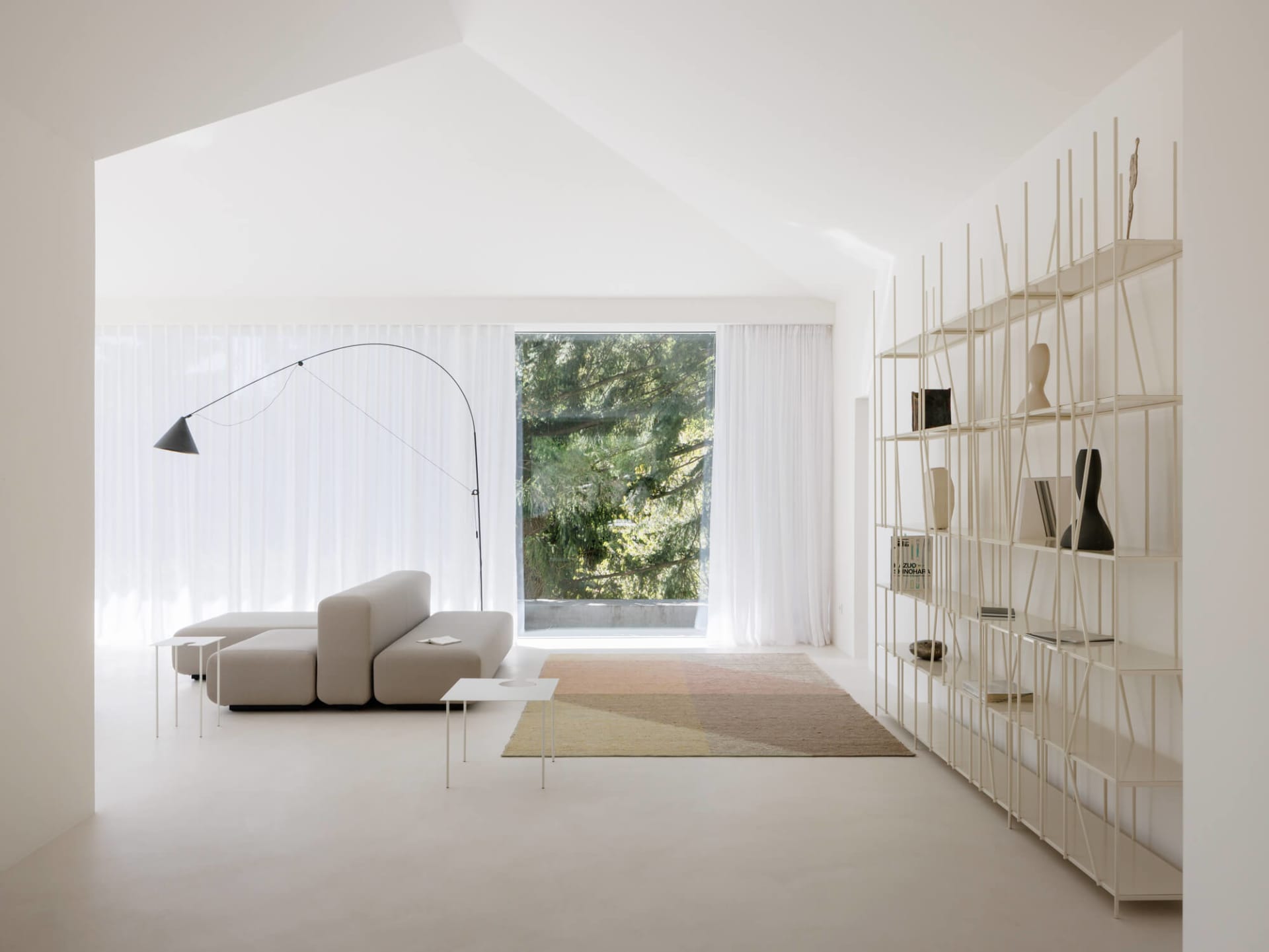 Bright minimalist living room featuring a beige modular sofa, colorful geometric area rug, tall white metal open shelving unit, and a black arc floor lamp in front of a large window with sheer curtains.