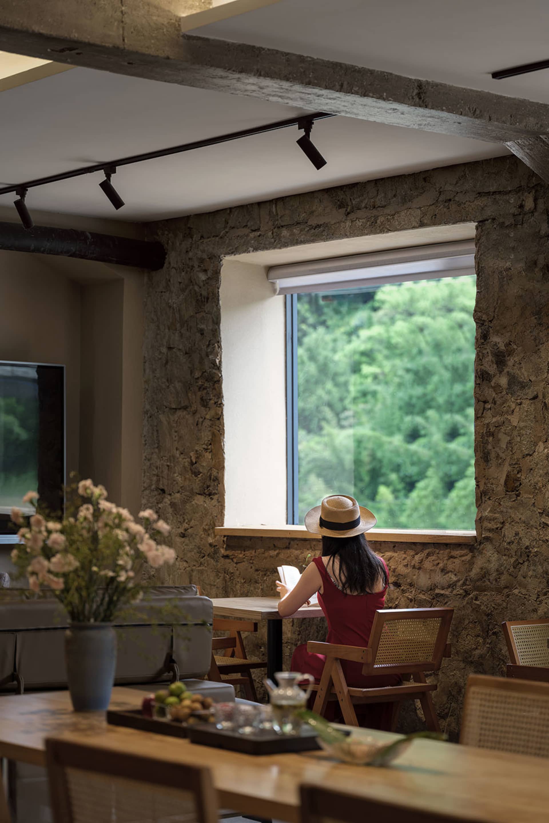 Woman sitting at a wooden dining table reading in front of a large window with a lush green forest view, surrounded by exposed stone walls and wooden furniture.