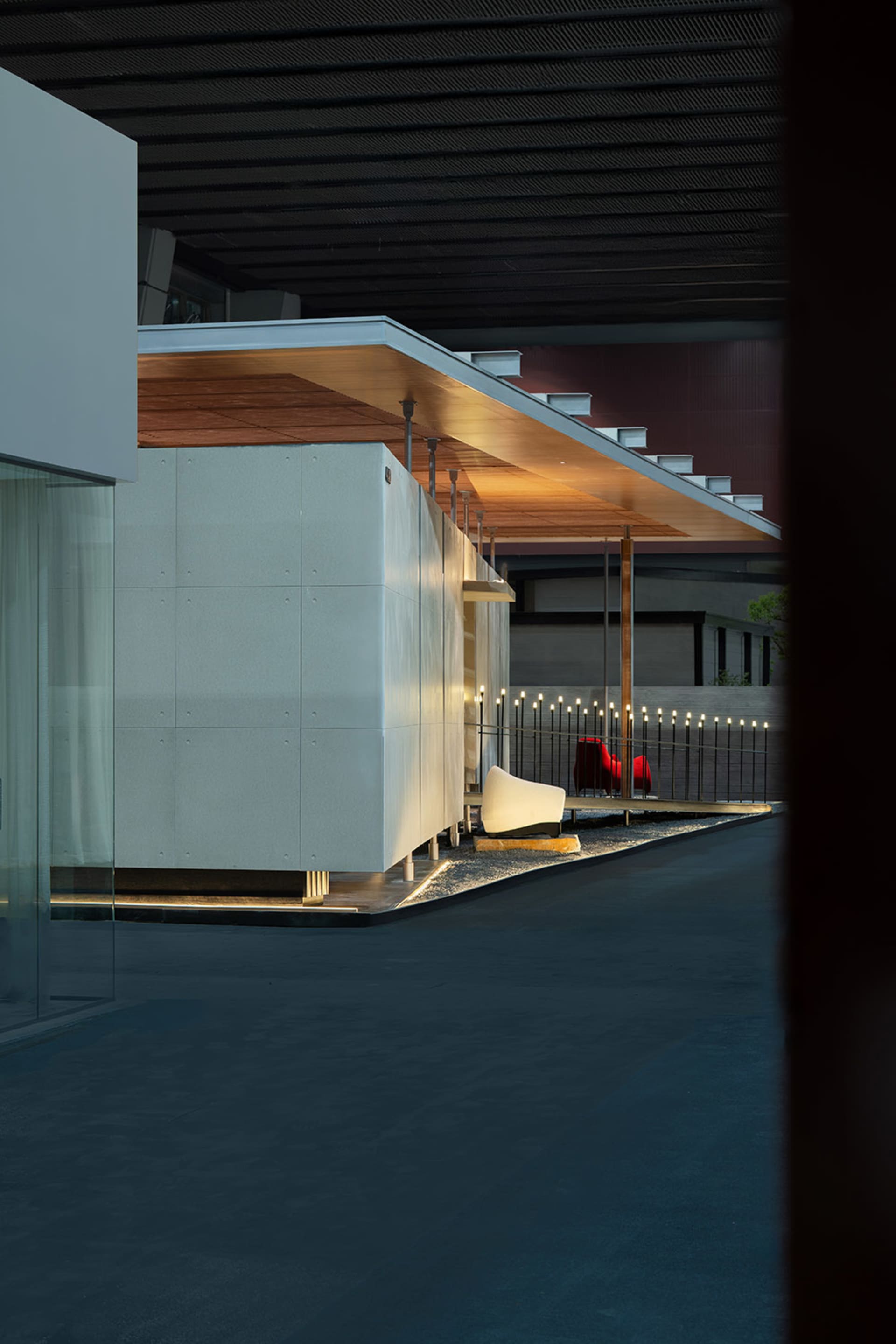 Modern minimalist courtyard at night featuring white concrete walls, a cantilevered wooden ceiling with integrated lighting, gravel garden, sculptural white bench, and red chairs in the background.