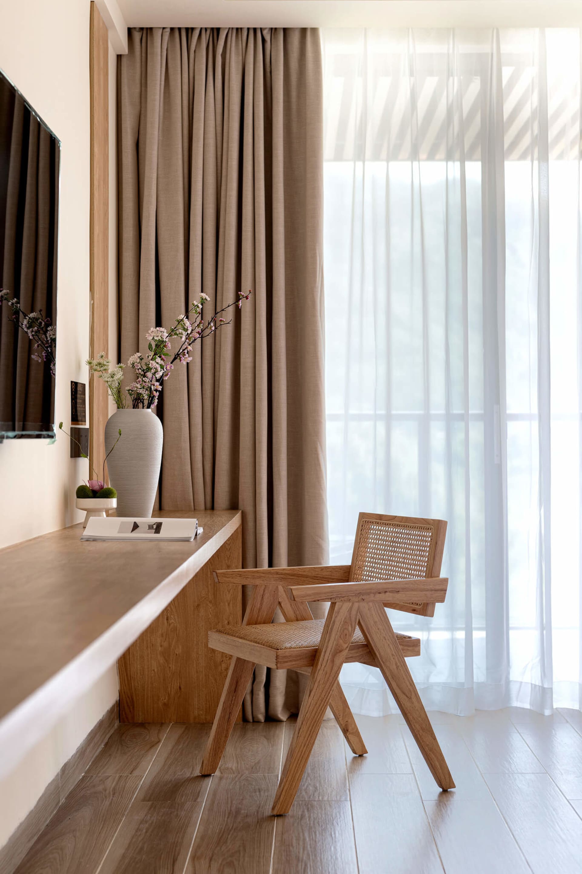 Minimalist wooden armchair with rattan seat and backrest placed next to a built-in oak desk and large window with sheer curtains