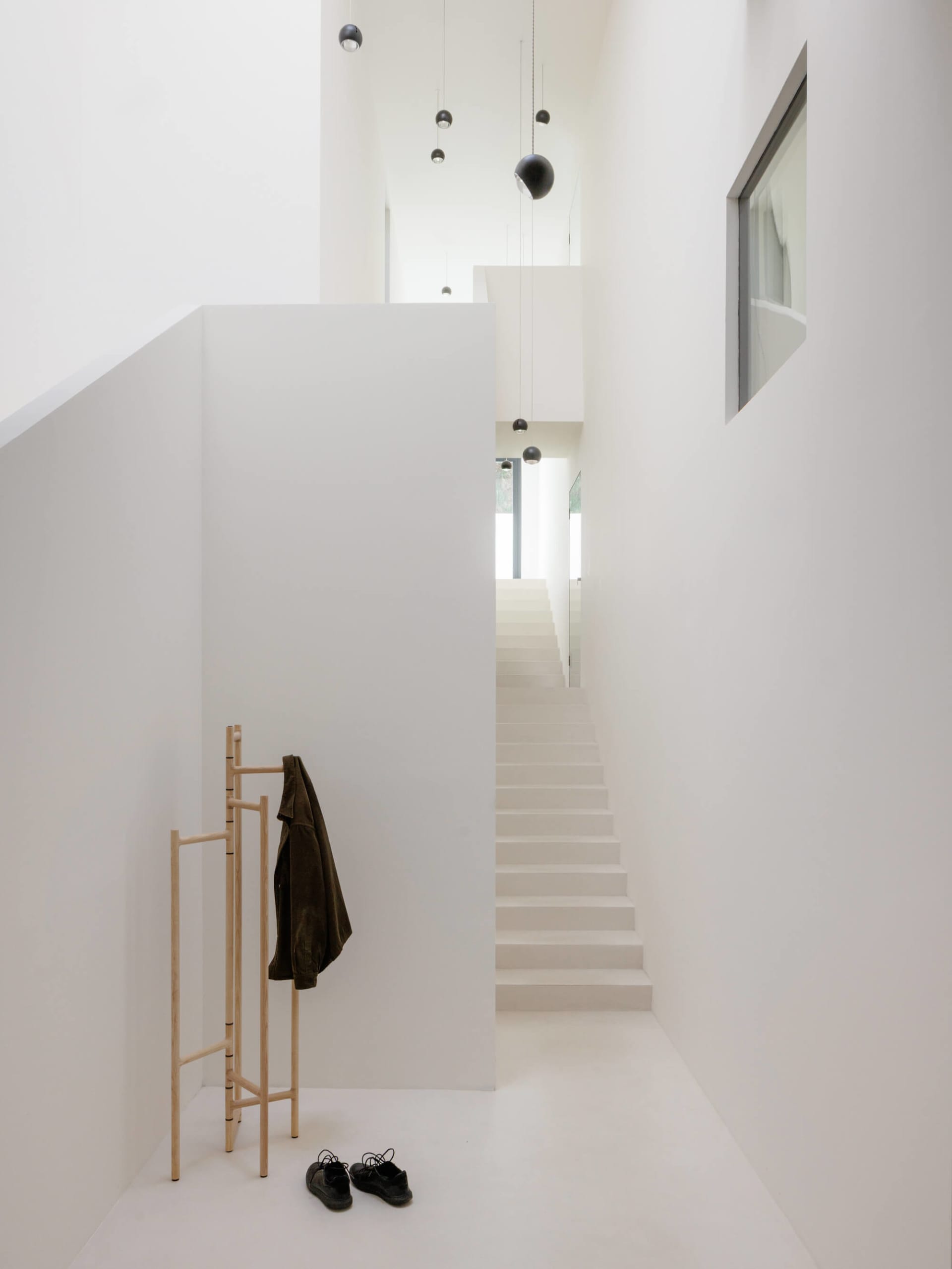 Minimalist white double-height entryway featuring a straight staircase, wooden coat rack with dark green jacket, black shoes on floor, and multiple black spherical pendant lights hanging from high ceiling.
