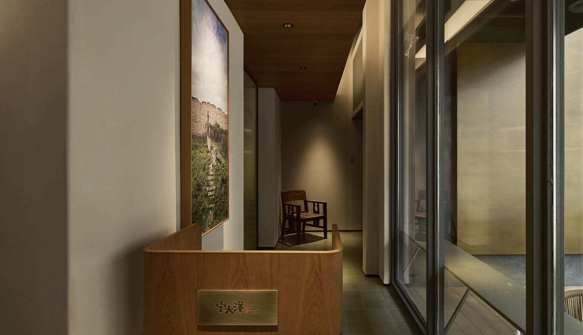 Narrow modern hallway featuring a curved oak reception desk, large landscape photograph, wooden chair, and warm ambient lighting.