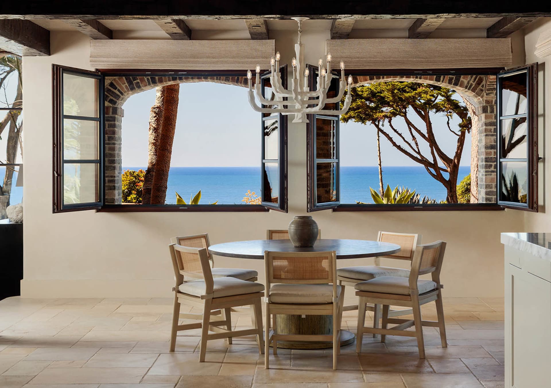 Round dining table with four cane-back chairs positioned in front of large arched windows opening to a bright blue ocean view, white chandelier hanging from beamed ceiling, limestone tile floor.