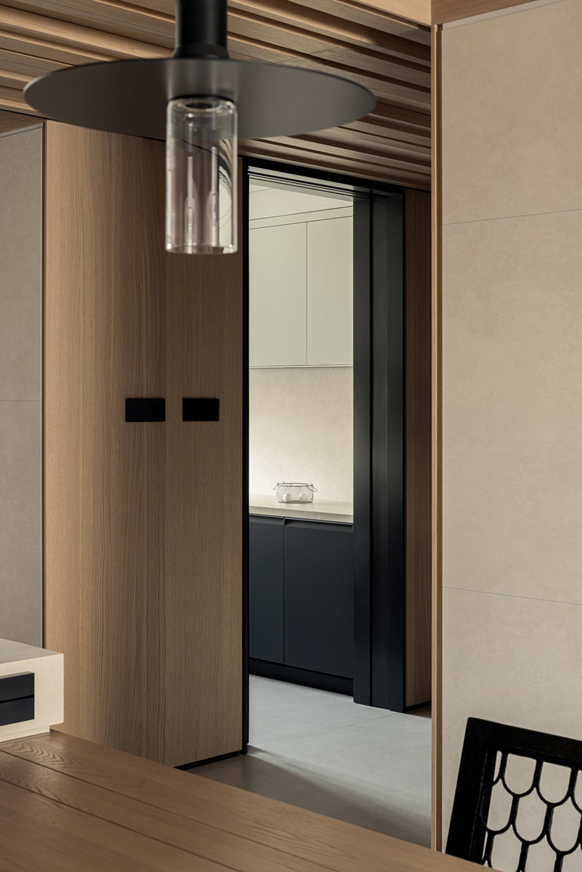 Modern minimalist dining area looking through a black-framed opening into a kitchen with warm oak cabinetry and matte black lower cabinets under soft lighting.