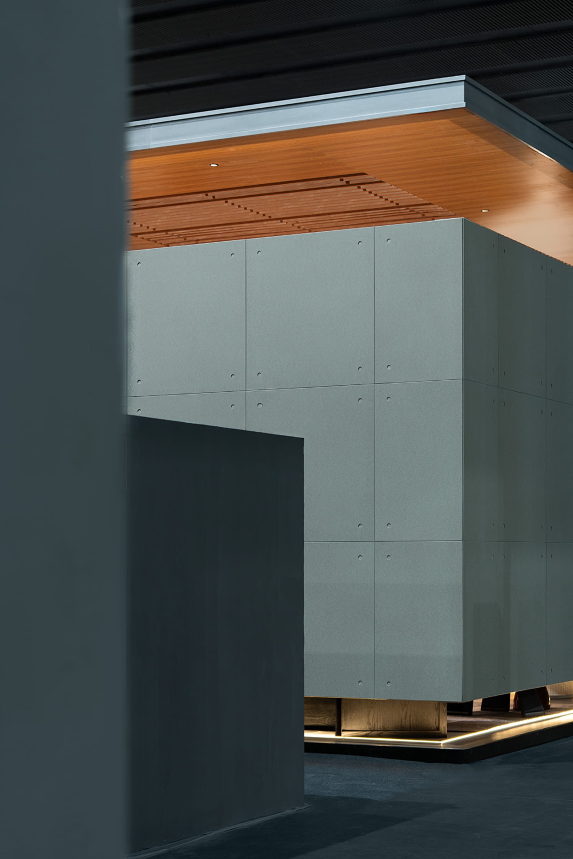 Modern architectural interior featuring a large floating concrete cabinet with visible bolt details, warm wood ceiling with linear slats, and dramatic indirect lighting at the base.