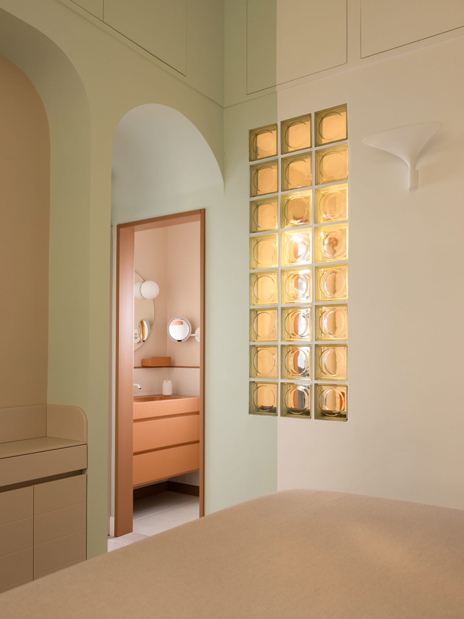 Modern minimalist bedroom corner featuring sage green walls, a peach vanity visible through an arched doorway, illuminated yellow glass block wall, and a built-in beige storage bench.