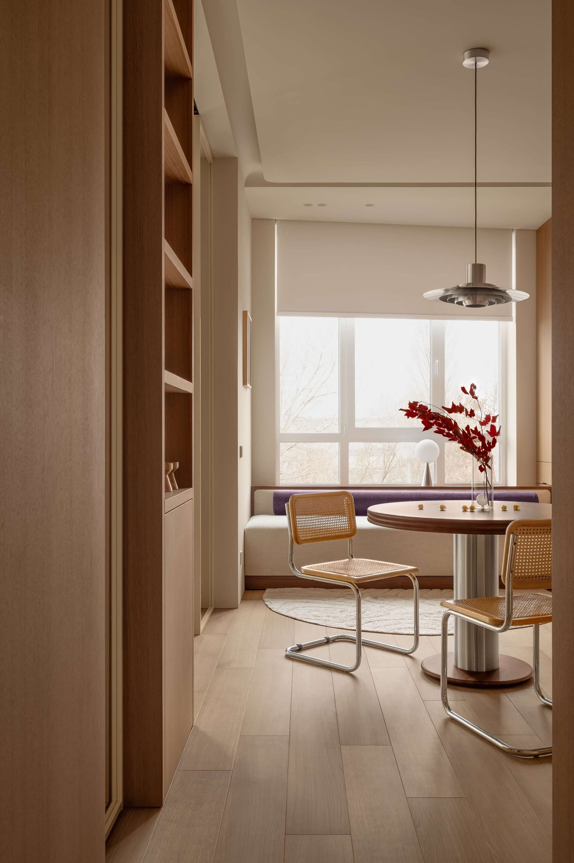 Modern minimalist dining nook with two cane and chrome chairs, round walnut table, built-in window seat with purple cushion, and large window overlooking trees.