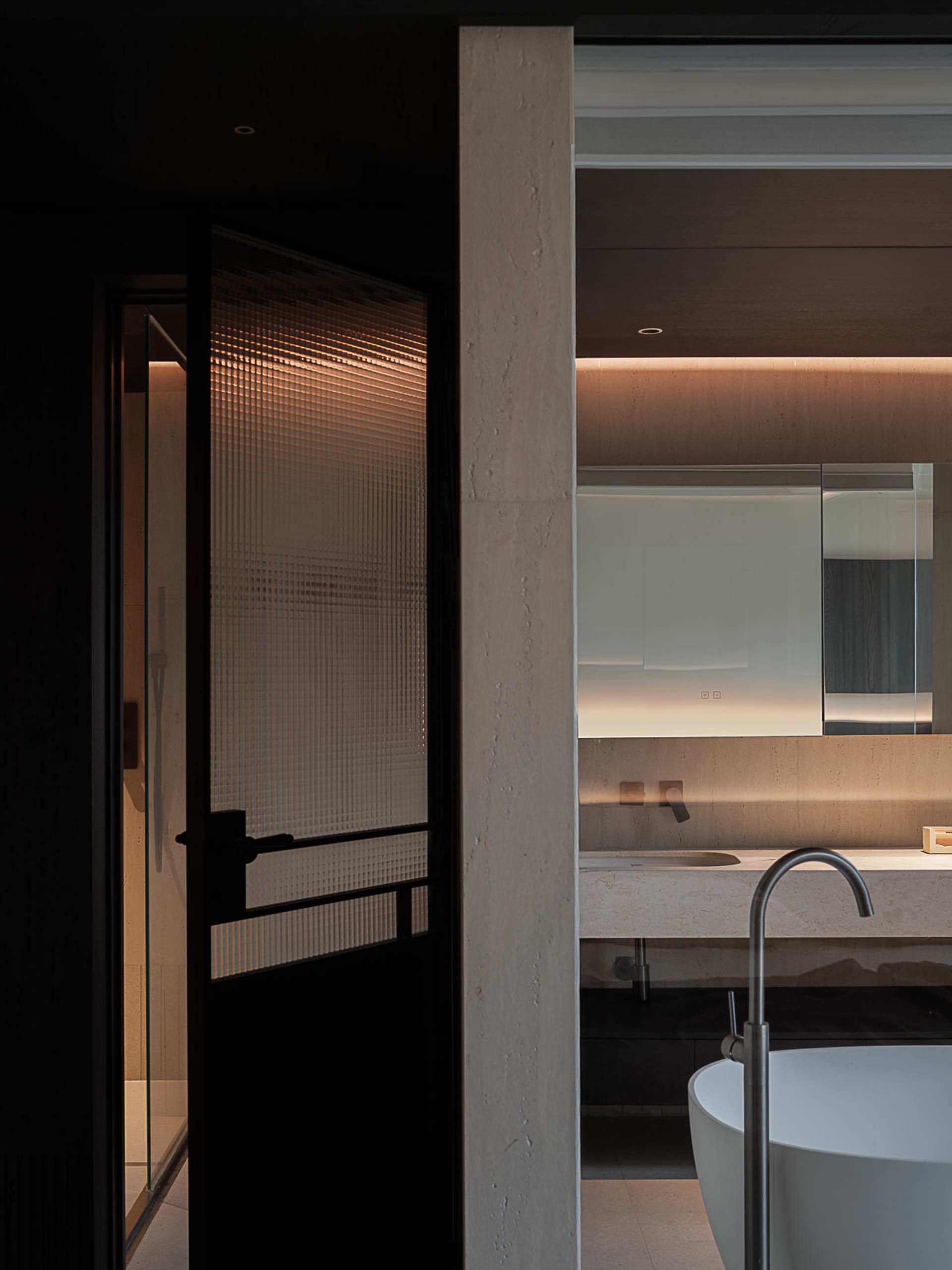 Modern minimalist bathroom featuring raw concrete walls, a white freestanding bathtub with a curved floor-mounted faucet, a stone floating vanity, and a black-framed ribbed glass door with warm backlighting.