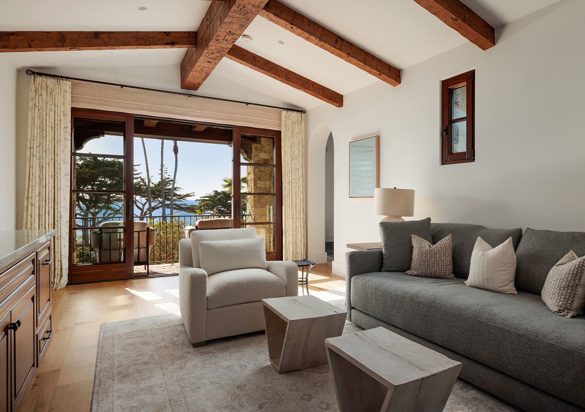 Bright coastal living room with white walls, reclaimed wood ceiling beams, grey sofa, cream armchair, wooden coffee tables, and large glass doors opening to an ocean view with palm trees.