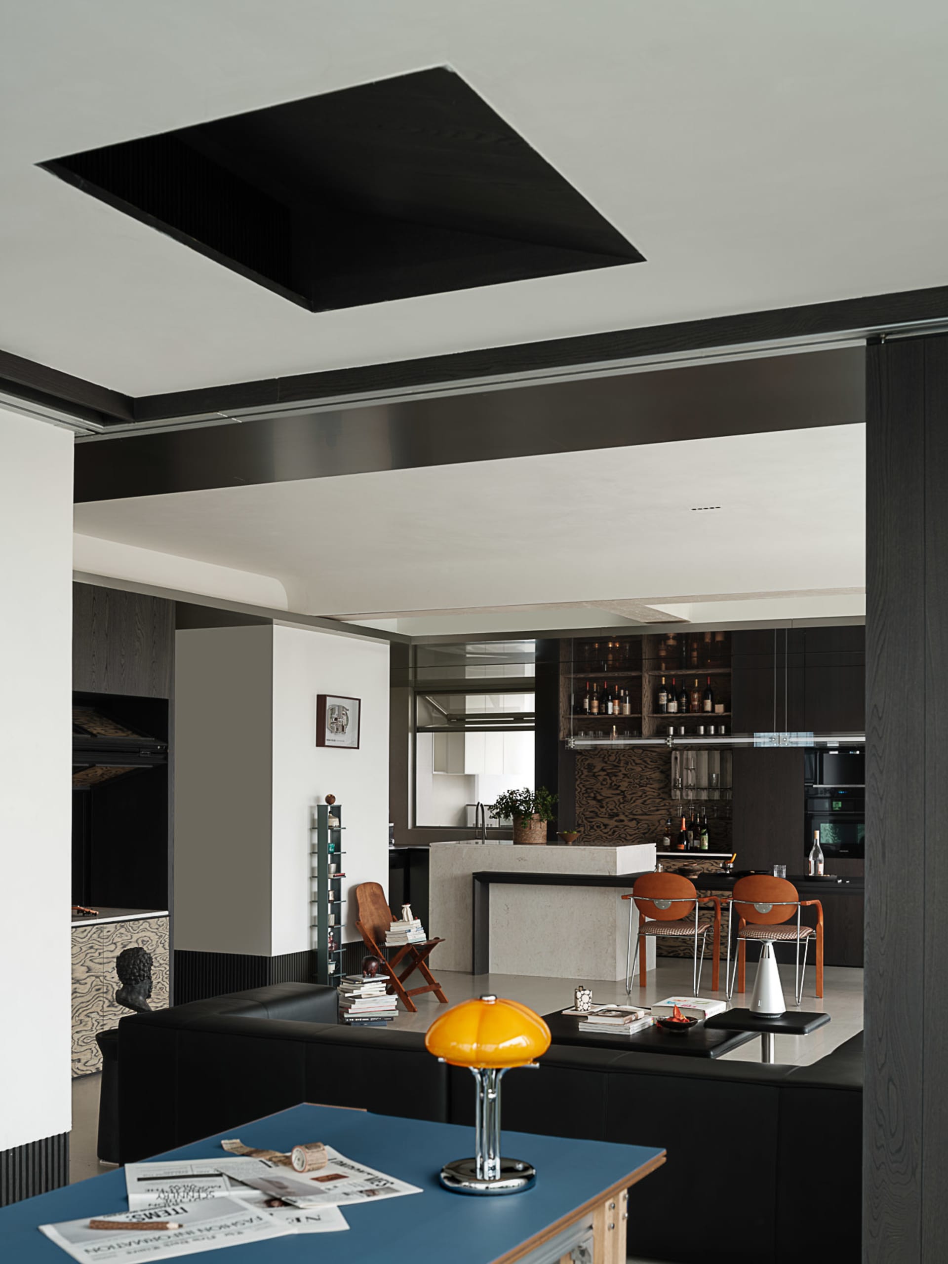 Modern open plan apartment interior featuring a black leather sofa, blue coffee table with yellow lamp, concrete kitchen island, burnt orange dining chairs, and dark wood architectural details.