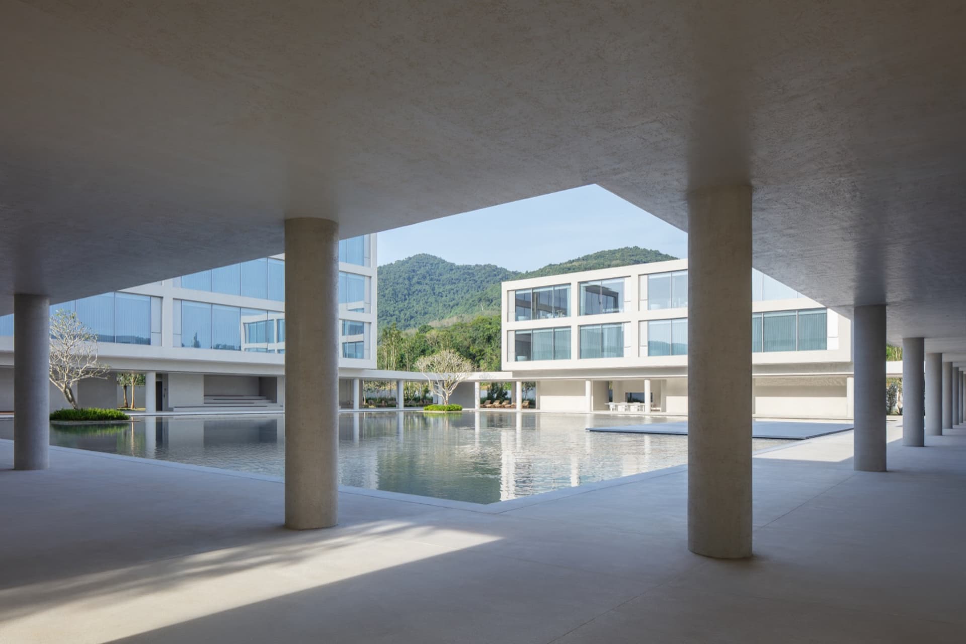 View from a shaded concrete colonnade overlooking a large rectangular reflecting pool, modern white buildings, and forested mountains in the background.