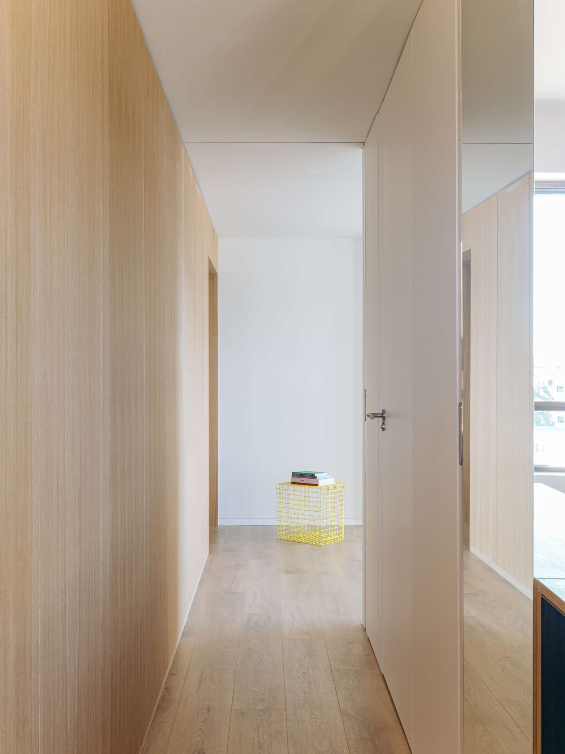 Narrow minimalist hallway lined with light oak wood paneling and cabinetry, featuring a bright yellow wireframe stool holding books at the end of the corridor.