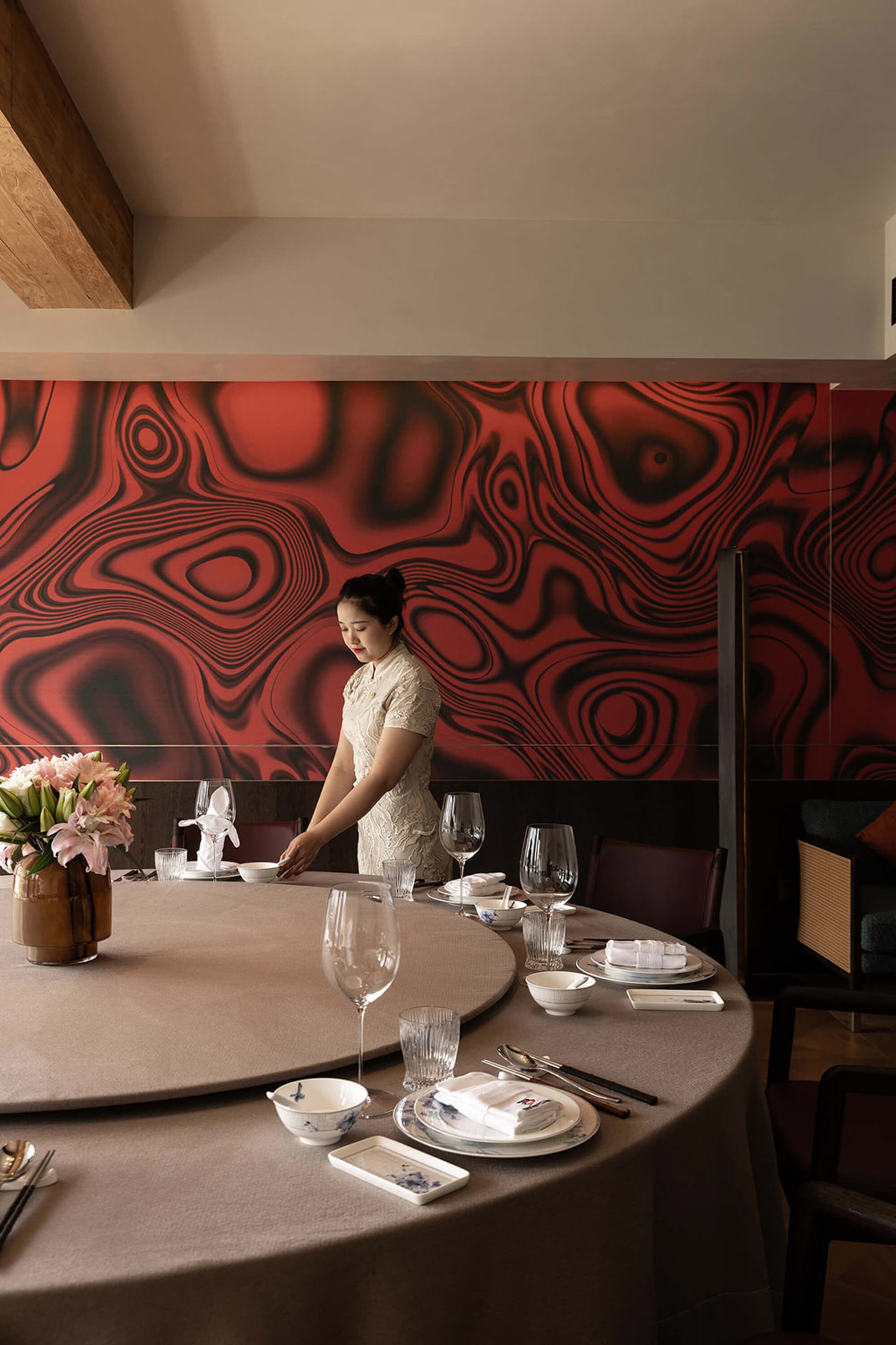 Elegant round dining table set with porcelain and glassware in a modern Chinese restaurant featuring a bold red and black swirling mural wall, with a female server in a white lace qipao adjusting the tableware.
