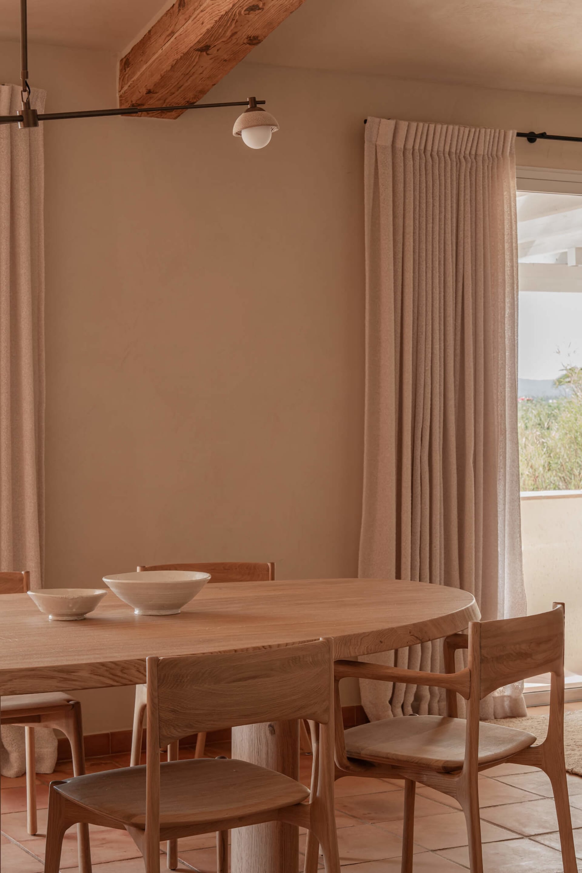 Warm minimalist dining room featuring a round oak dining table, matching oak chairs, exposed wooden ceiling beams, terracotta floor tiles, and beige linen curtains with a view to the outdoors.
