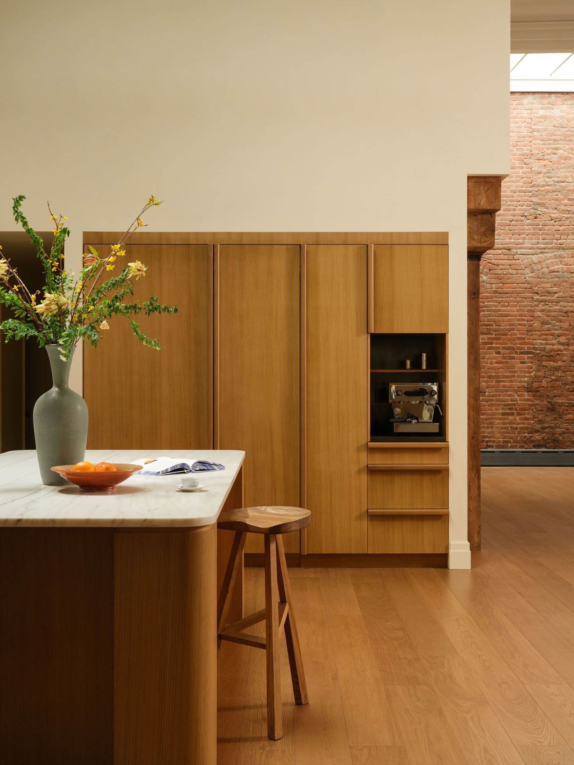 Modern minimalist kitchen featuring a curved oak island with white marble countertop, wooden stool, built-in oak cabinetry, and a large grey vase with yellow forsythia branches.